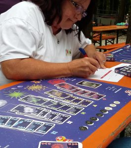 Bild: Spielefeld mit Karten sowie eine Frau, die Spielergebnisse auf Block notiert