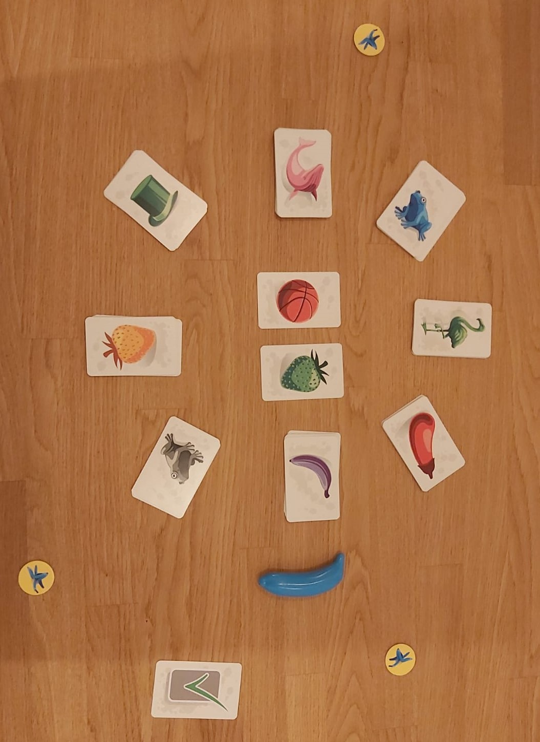 Zu sehen ist der Spielaufbau von Blue Banana: 2 Aufgabenkarten liegen in der Mitte umgeben von 8 gleich großen Stapeln mit Motivkarten. Außerdem hat jeder einen Strafchip zu Beginn vor sich liegen.