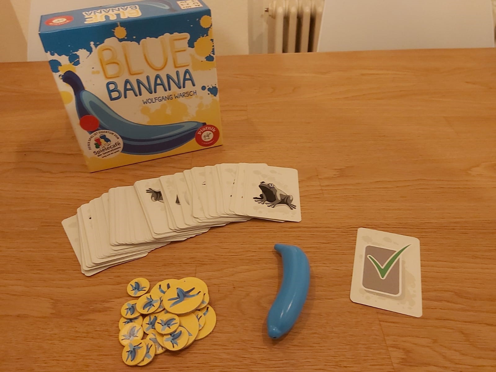 Auf einem Holztisch stehen die Verpackung von dem Spiel Blue Banana, sowie das gesamte Material bestehend aus den Motivkarten, einer blauen Plastikbanane, den gelben Strafchips und der Aufgabenkarte.