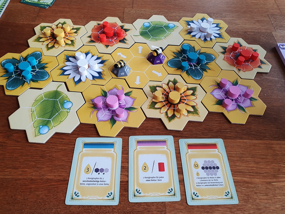 Aufgebauter Spielplan aus Blütenplättchen, darauf Spielsteine und davor Aufgabenkarten