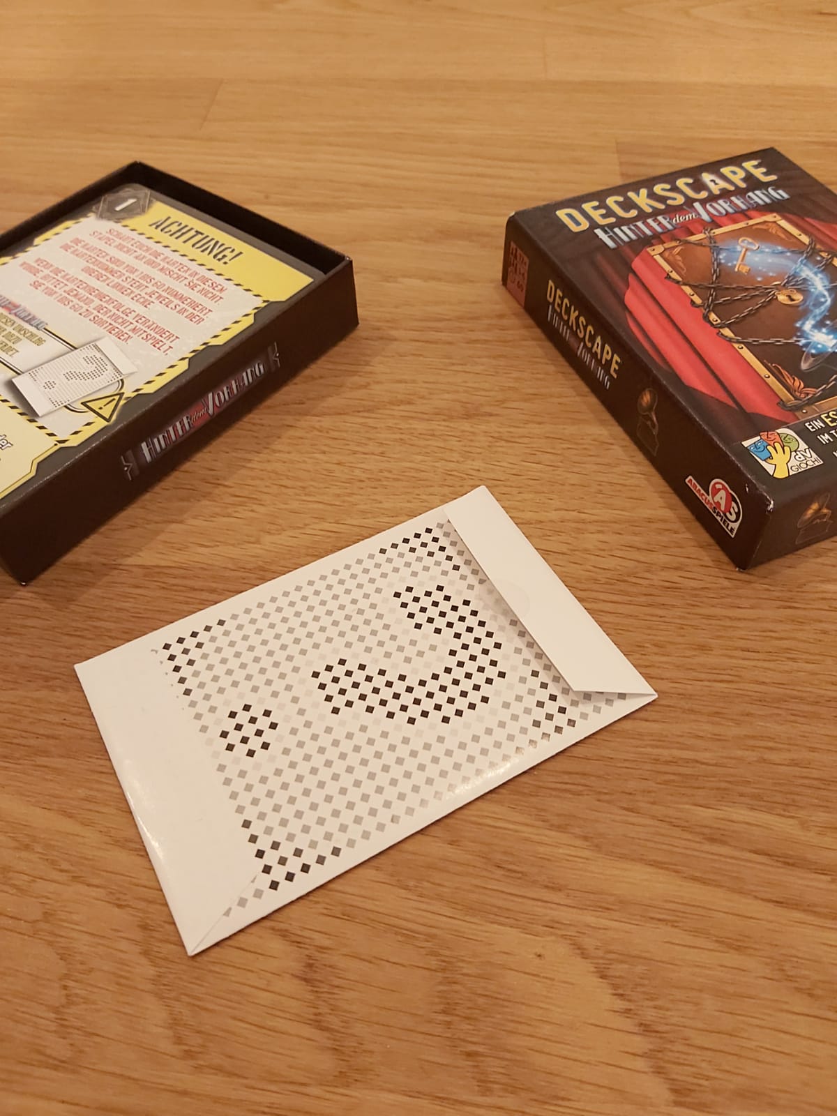 Zu sehen ist die Verpackung sowie Spielmaterial von Deckscape - hinter dem Vorhang