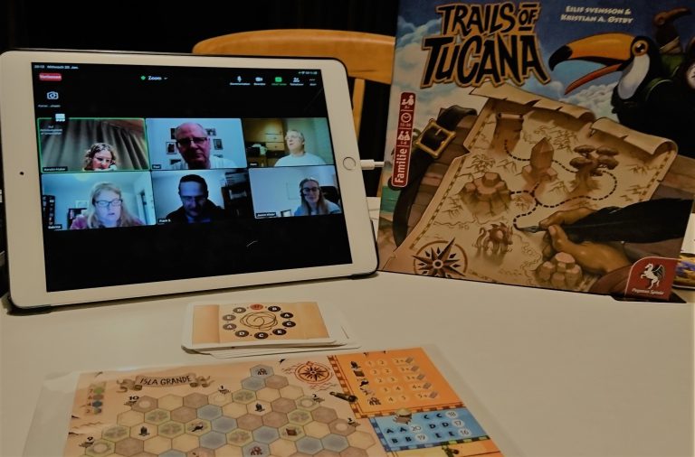 Fotografie einer Online Spielerunde, Links ein Tablet welches die Zoomsitzung zeigt, rechts davon der Spielkarten von Trails of Tuscana, vor beiden Gegenständen ist das Spiel aufgebaut