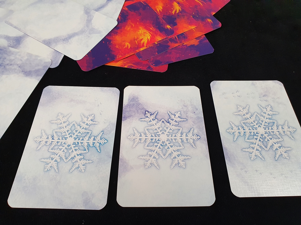 Drei Karten mit Schneekristallen liegen vor dem fast zugedeckten Feuerfeld