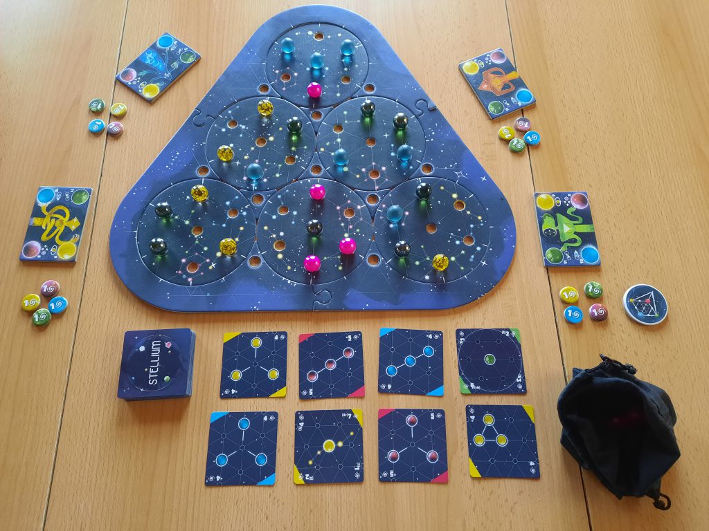 Das aufgebaute Spielbrett sieht wie ein Dreieck aus mit runden Scheiben in der Mitte und Löchern für die Murmeln), verschiedenfarbige Murmeln auf dem Spielbrett verteilt, acht aufgedeckte Aufgabenkarten liegen unter dem Spielbrett, ein Stapel umgedrehter Aufgabenkarten liegt daneben, Spielmaterial für 4 Spieler liegt verteilt neben dem Spielbrett, ein Beutel mit Murmel liegt bereit
