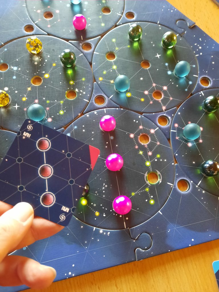 Teile des Spielbretts sind zu sehen, drei rote Murmeln liegen in einer Reihe nebeneinander, daneben ist die Aufgabenkarte zu sehen