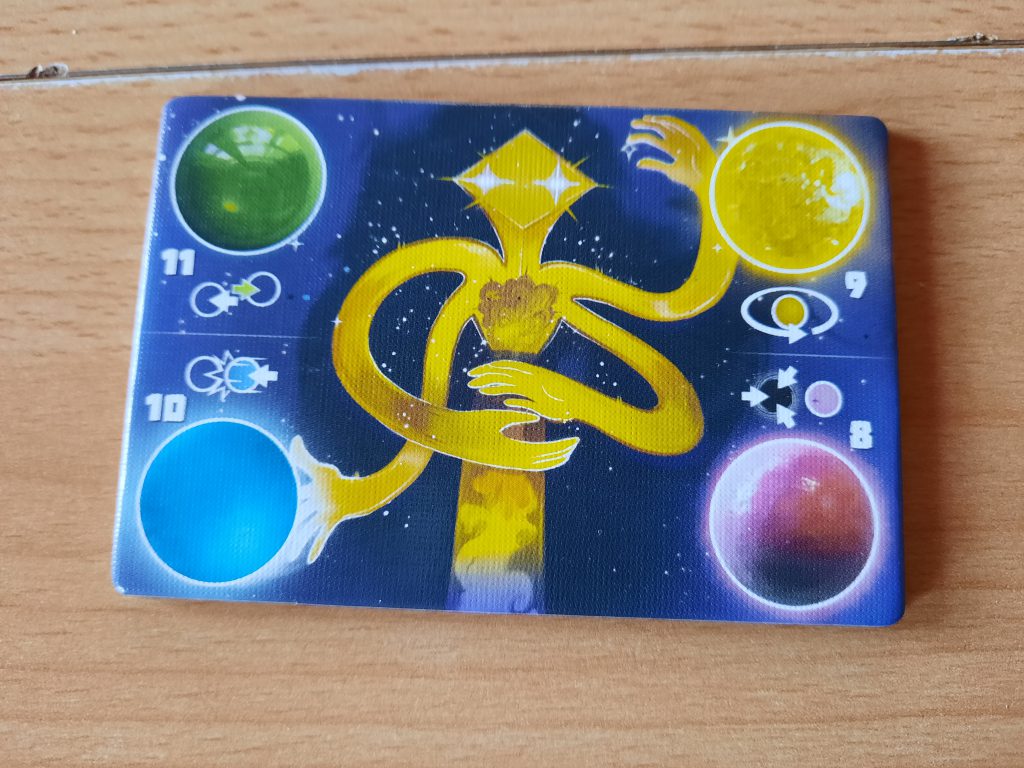 eine bunte viereckige Karte, das Spielhilfeplättchen ist zu sehen, auf dem Plättchen sind in jeder Ecke eine andersfarbige Murmel und darunter die Erklärungen abgebildet