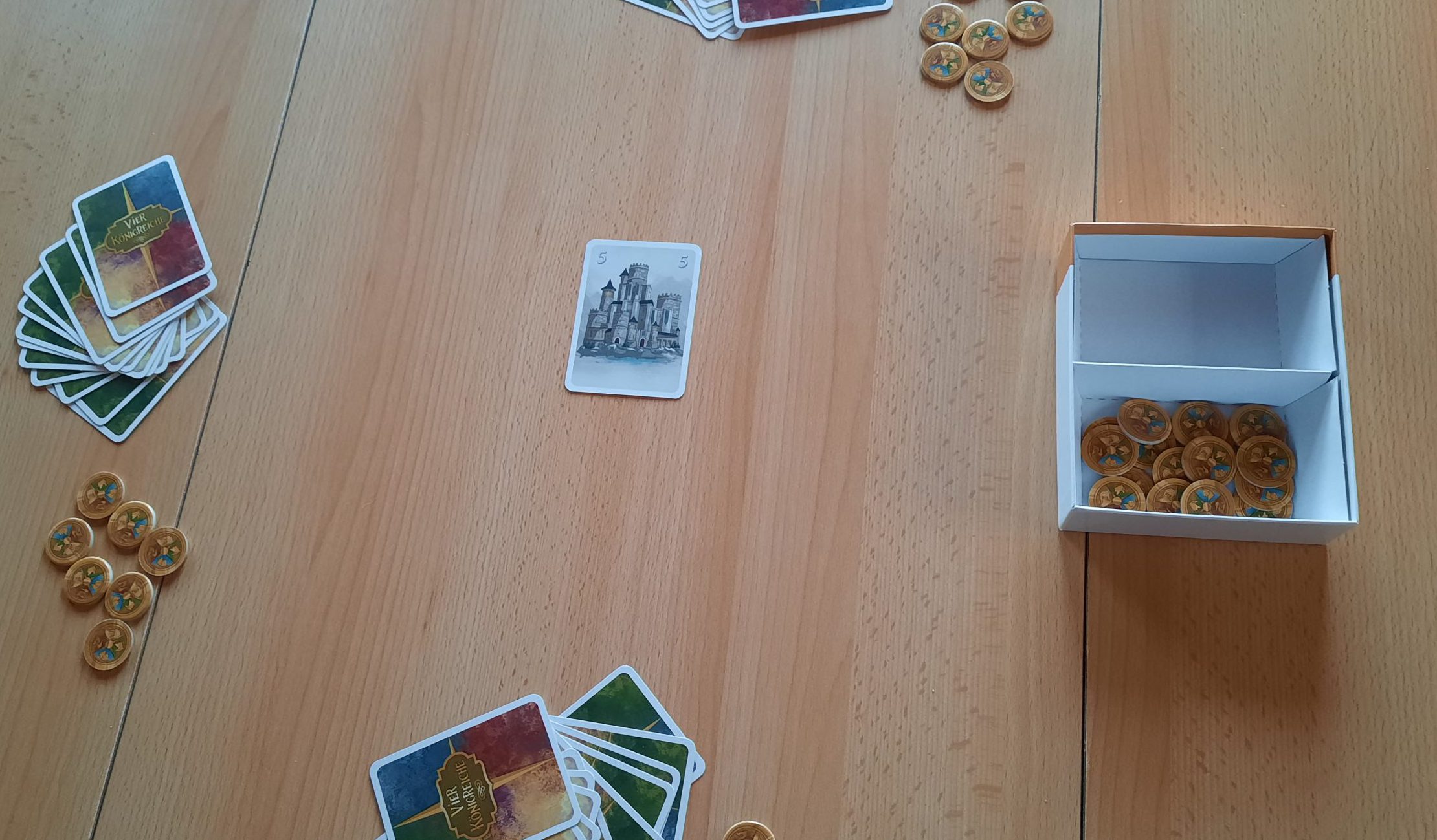 Die Karte mit der blauen 5 liegt offenen in der Tischmitte. Drumherum liegen die Handkarten der Spielenden (verdeckt) und ihre Münzen.