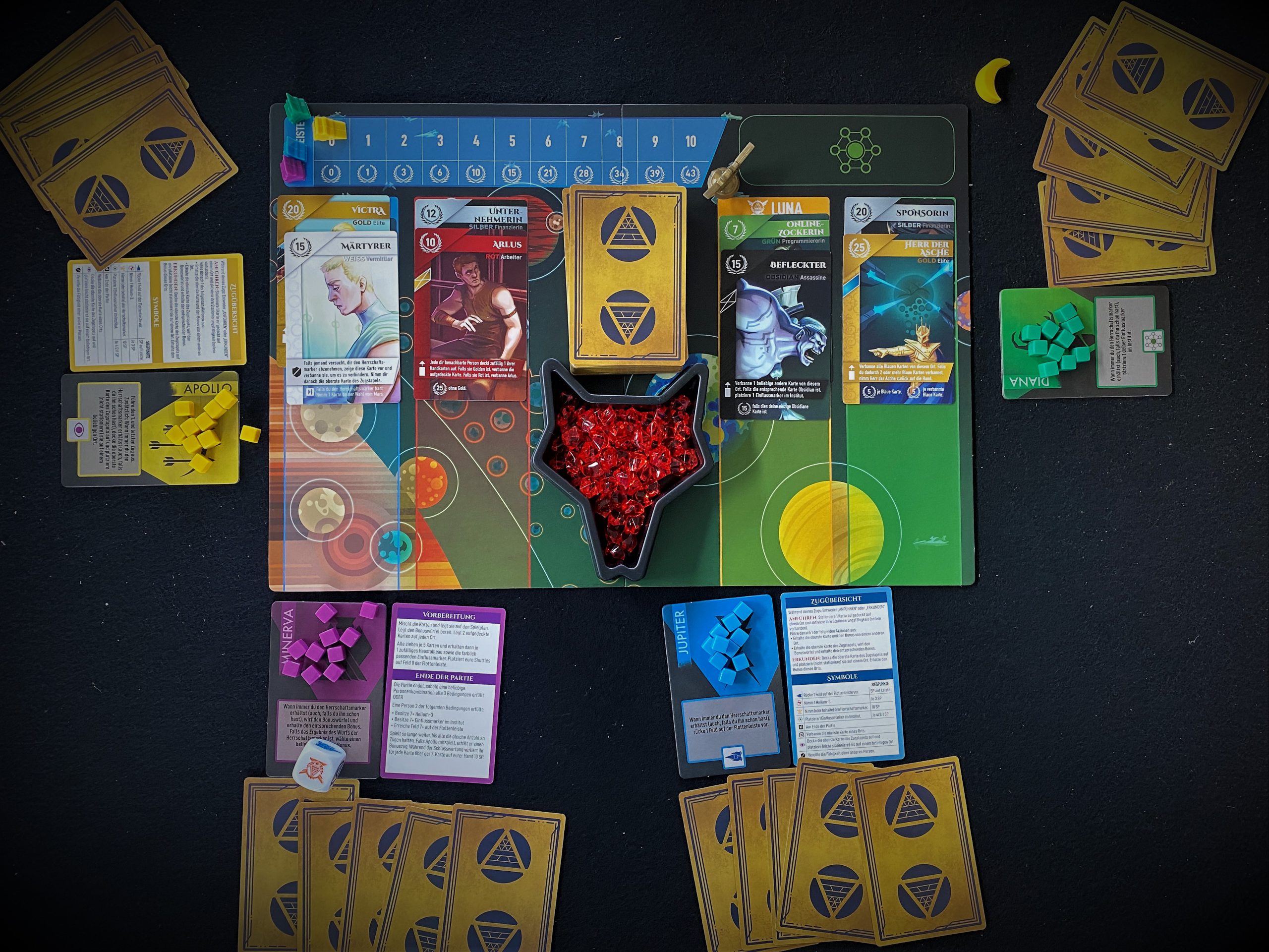 Zentral liegt das Spielbrett, außen herum legen die Spielenden das Material ab.