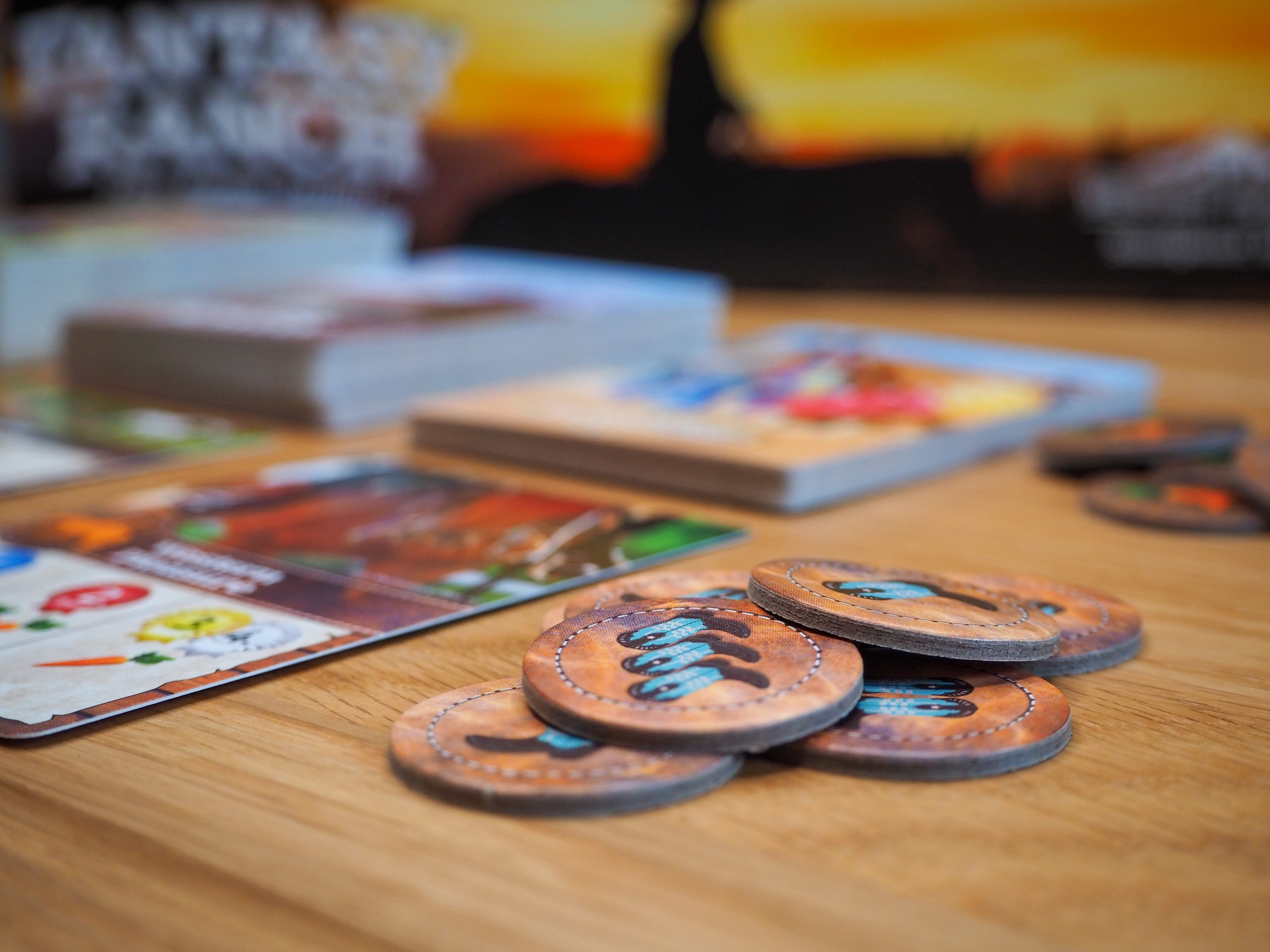 Im Vordergrund erstrahlt ein zusammengewürfelter Haufen Ausrüstungsplättchen in warmem Licht. Im Hintergrund sind unscharf einige Kartenstapel und ein Teil der Spielepackung zu sehen.