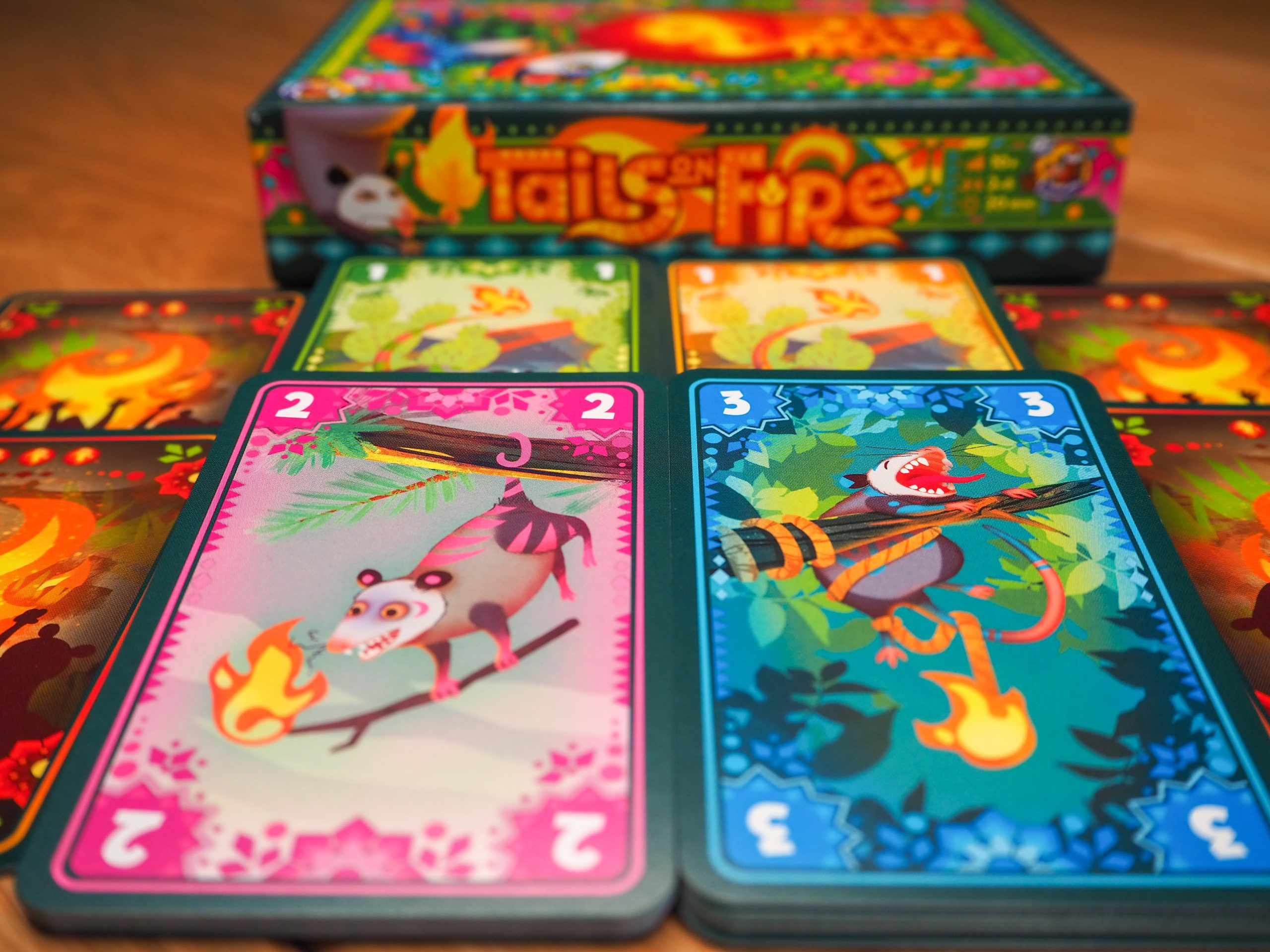 Mittig im Hintergrund ist verschwommen die Spielschachtel von der langen Seite zu erkennen. Davor liegen die Stapel der verschiedenen Farben des Opossum- Karten.