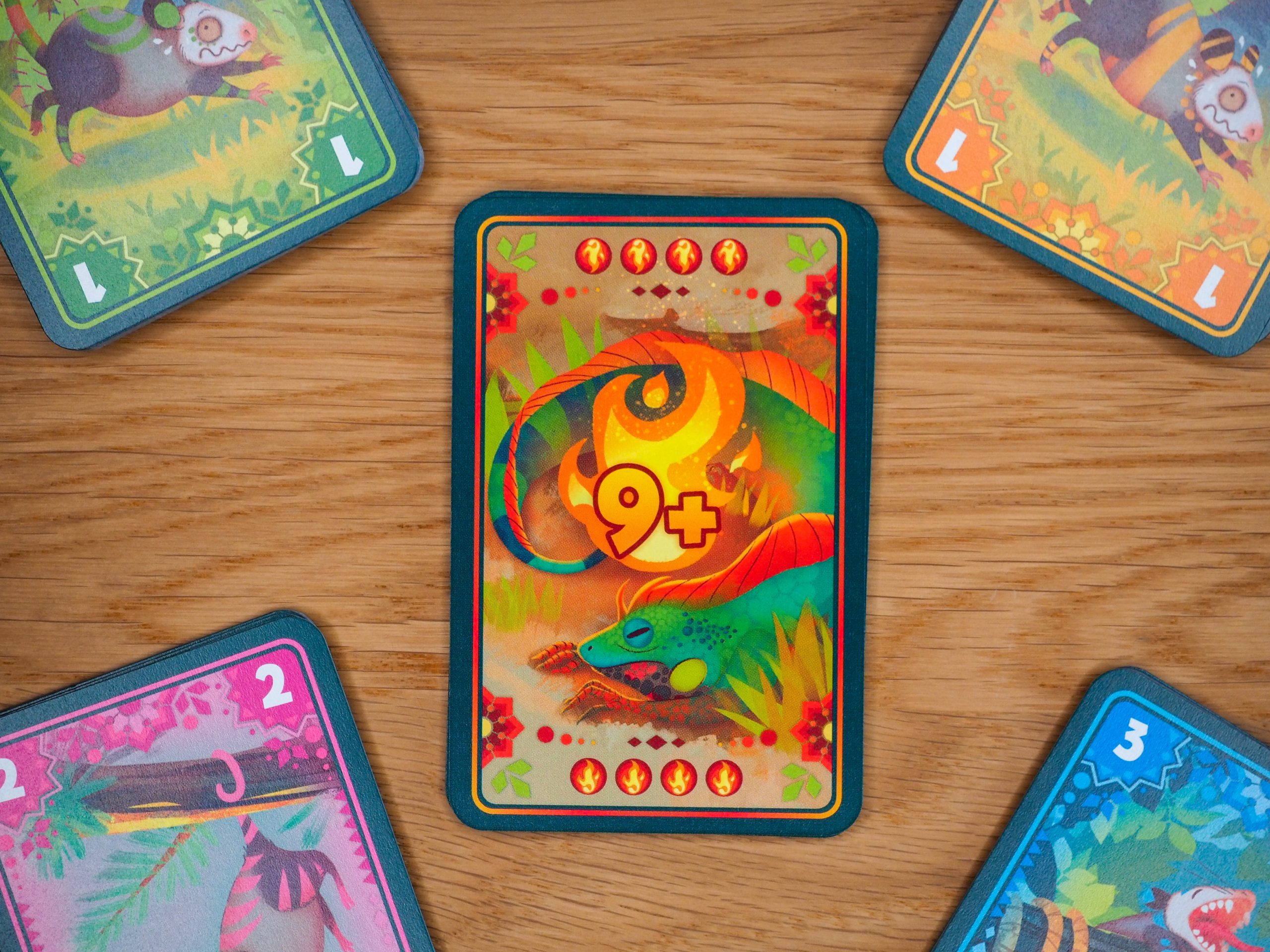 Zentral liegt die Feuerkarte des Iguana-Stapels mit der Zahl 9+ in einem leuchtenden Rot. Kreisförmig um die Feuerkarte herum sind die Stapel der verschiedenen Mitspielenden platziert. Sie haben die Farben grün, orange, blau und pink.