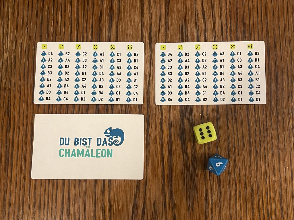Auf dem Bild sind 3 Karten abgebildet, 2 von denen zeigen die Codekarten an und eine Karte trägt nur den Schriftzug "Du bist das Chamäleon". Auf der rechten Seite unten sind außerdem ein gelber sechsseitiger Würfel und blauer achtseitiger Würfel abgebildet.