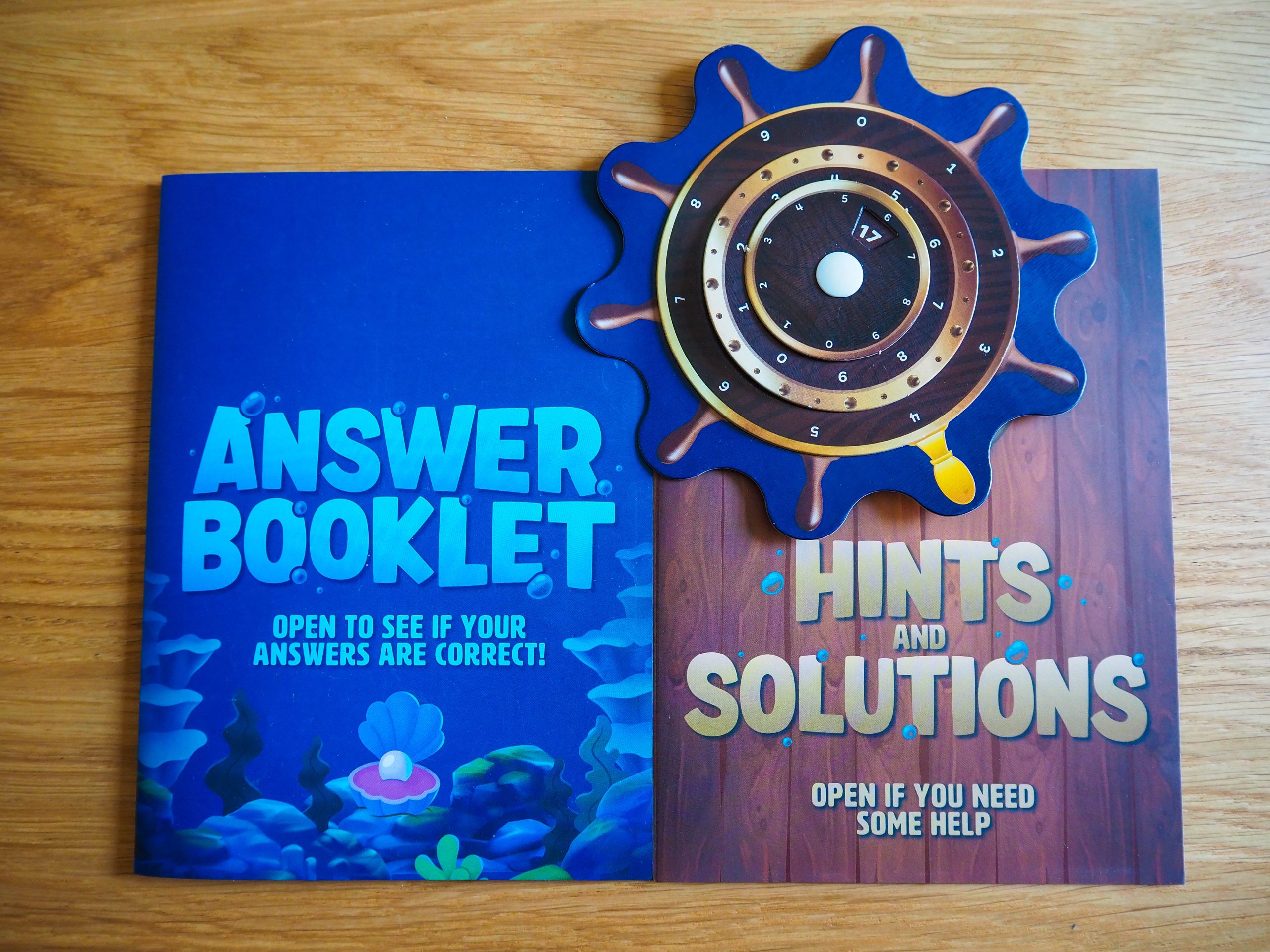 Links im Bild ist eine Broschüre zu sehen, in der die Lösungen versteckt sind. Im unteren drittel der Broschüre erkennt ihr eine geöffnete Muschel mit Perle. Rechts daneben befindet sich die Broschüre mit Hinweisen und Lösungen. Ihr Hintergrund besteht aus braunen Holzdielen. Im oberen Drittel auf dieser Broschüre liegt das Steuerrad. Es ist rund herum mit verschiedenen Zahlen beschriftet.