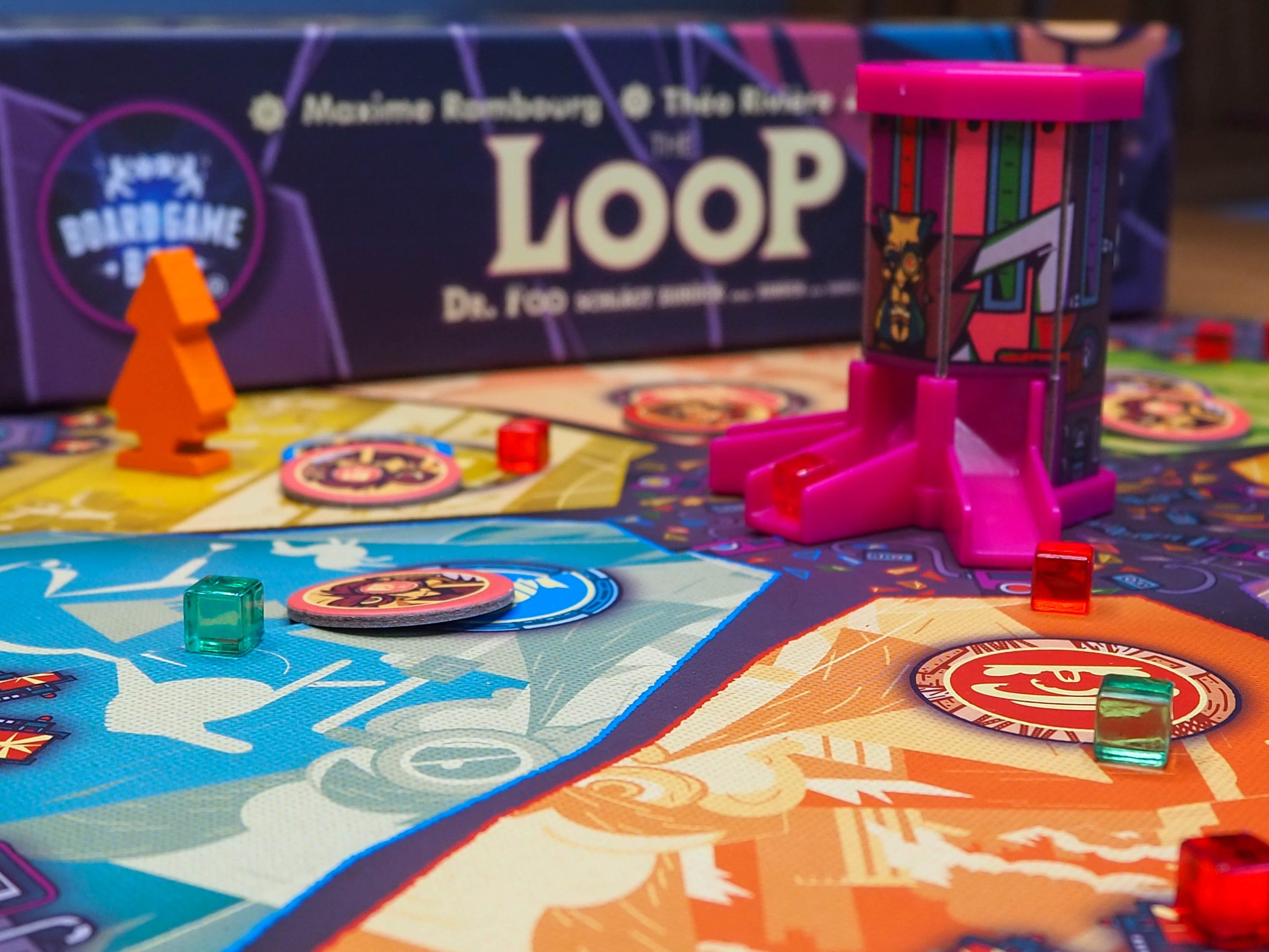 Verschwommen im Hintergrund erscheint die Seitenansicht der Spieleschachtel. Im Vordergrund das Spielbrett mit der 3D Zeitmaschine. Diese ist ein Turm mit drei verschiedenen Ausgängen. An den Ausgängen liegen Zeitrisswürfel. Außerdem ist im Hintergrund verschwommen eine Spielfigur in orange zu sehen und verschiedene Duplikate.