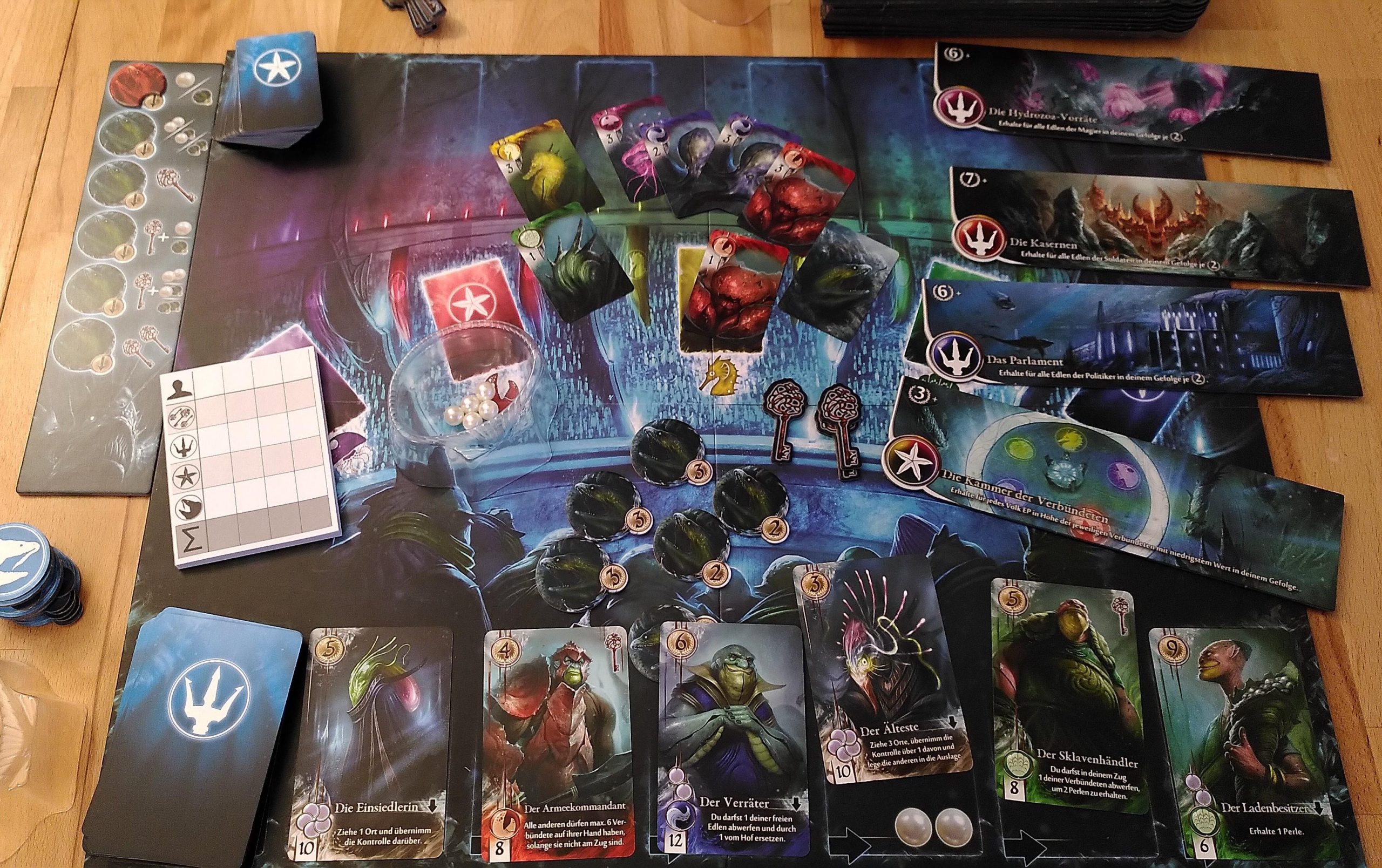 Überblick über das Spielmaterial und das Design von Abyss. Viele Karten mit Unterwasserwesen, Papp-Schlüssel und -Abzeichen, durchsichtige Muschelschalen mit Plastikperlen.