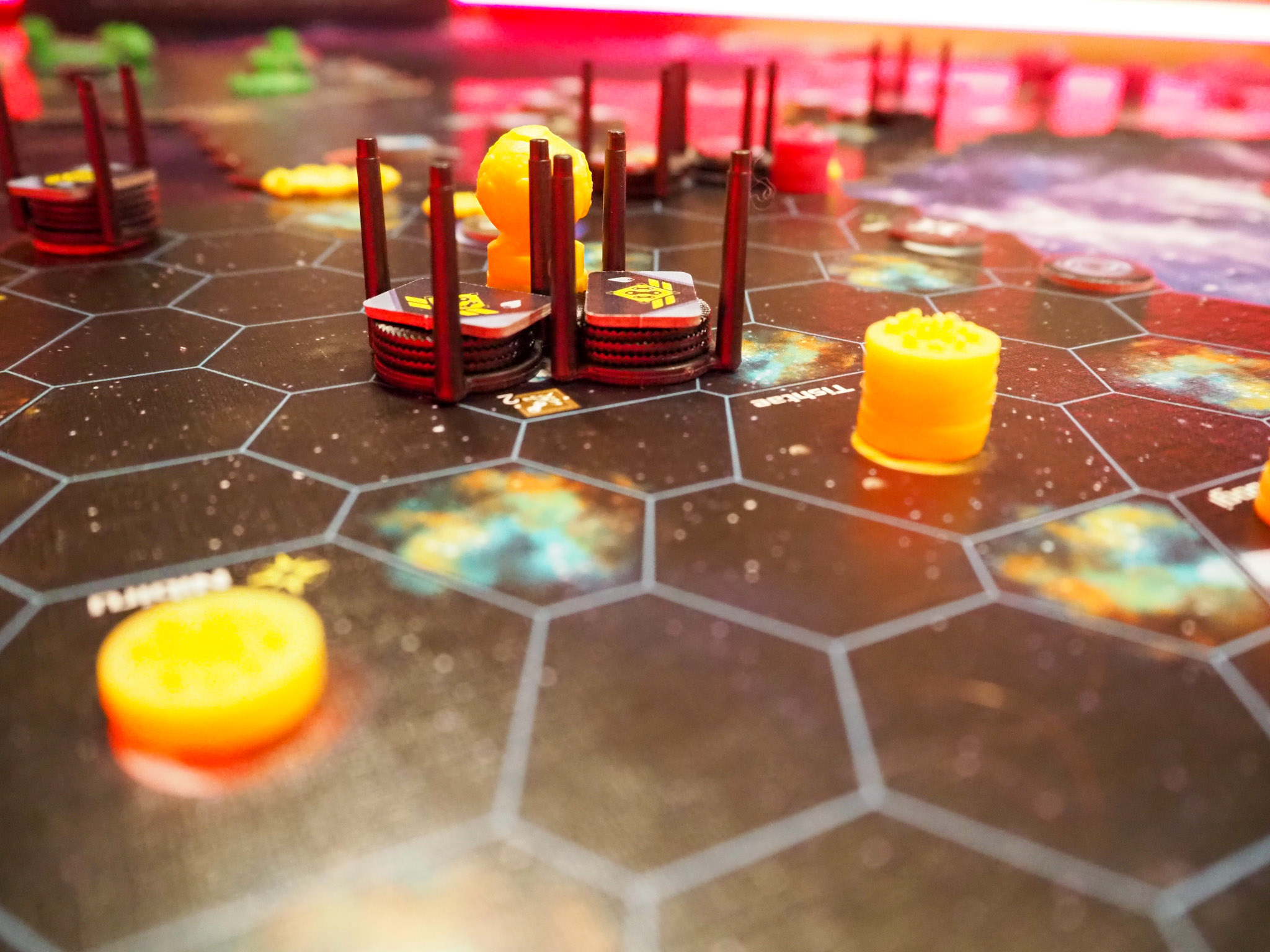 Das Bild zeigt den Spielplan, wobei der vordere und hintere Teil unscharf sind, während der mittlere Bereich scharf und deutlich zu sehen ist. In der Mitte befinden sich Flotten in ihren schwarzen Ständern. Der Hintergrund ist in einem hellen Pink gehalten, und es sind teilweise gelbe Bevölkerungsscheiben zu erkennen.