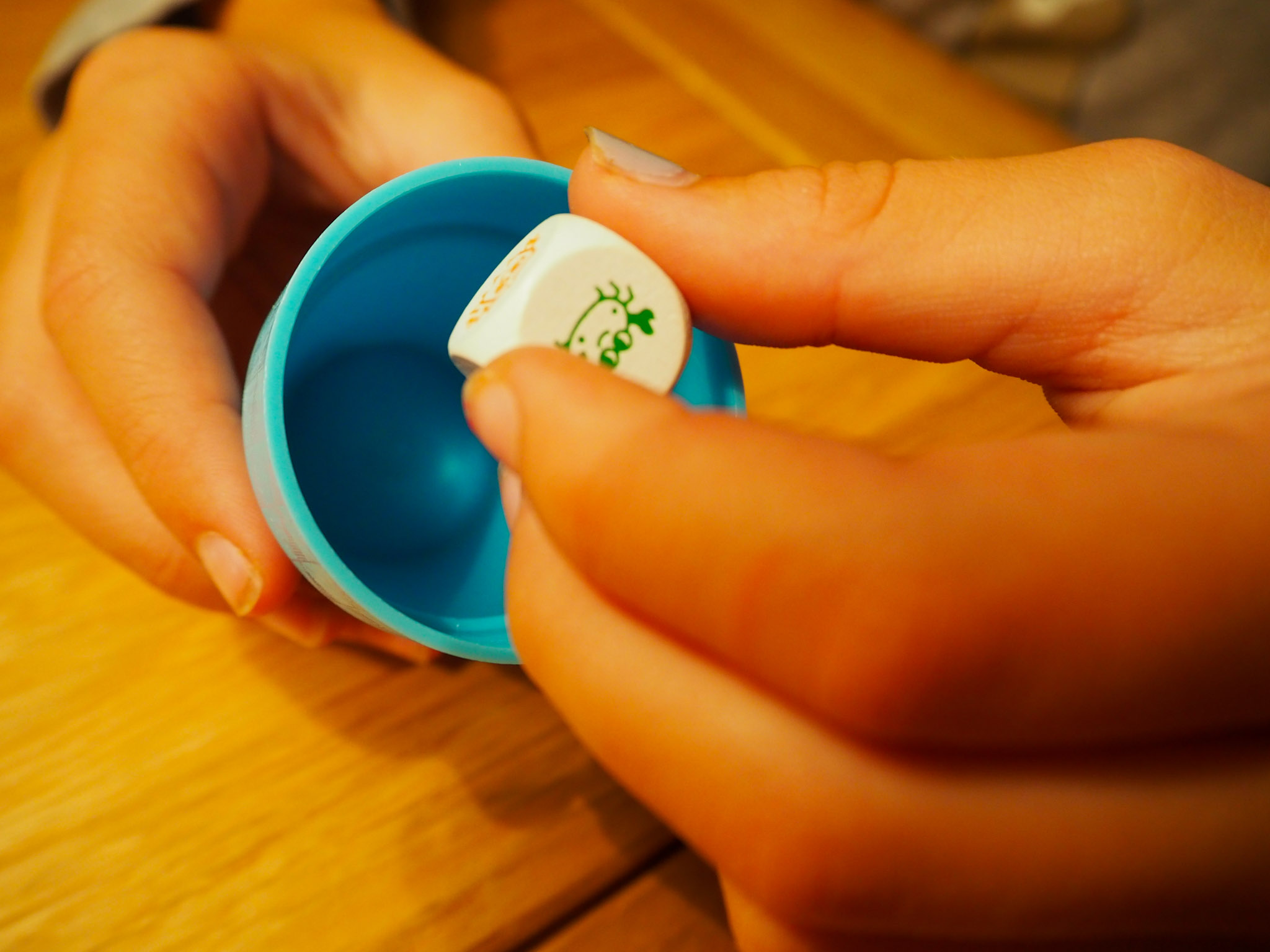 Eine Kinderhand hält einen Würfel zwischen dem Daumen und dem Zeigefinger und ist kurz davor ihn in den Würfelbecher, den es in der anderen Hand hält, hineinzuwerfen.