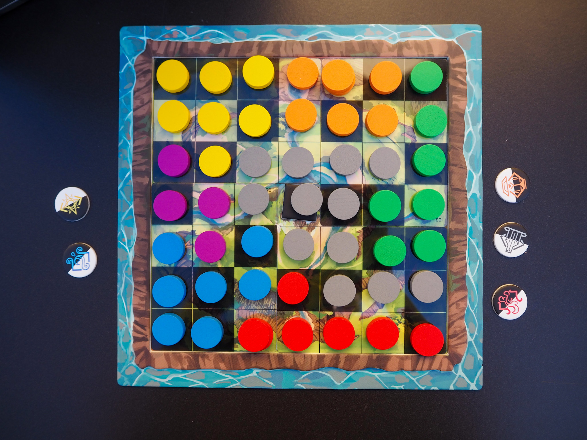 Auf diesem Bild wurde das Spielbrett von oben fotografiert. Die einzelnen Kästchen des Bretts sind voll mit verschiedenfarbigen Spielsteinen. Rechts neben dem Spielbrett liegen drei Siegplättchen und links davon zwei.