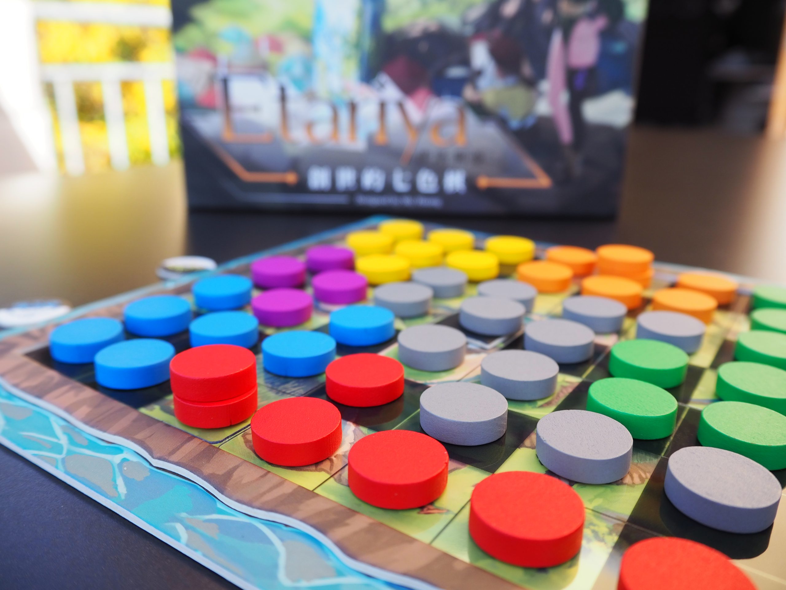 Der Vordergrund zeigt das Spielbrett. Auf jedem Feld liegt mindestens ein Spielplättchen in unterschiedlichen Farben. Vorne dominiert eher rot, wohingegen unscharf im hinteren Bereich die Farben gelb und orange dominieren. Im Hintergrund des Spielbretts erscheint die Spielschachtel von Etariya verschwommen.