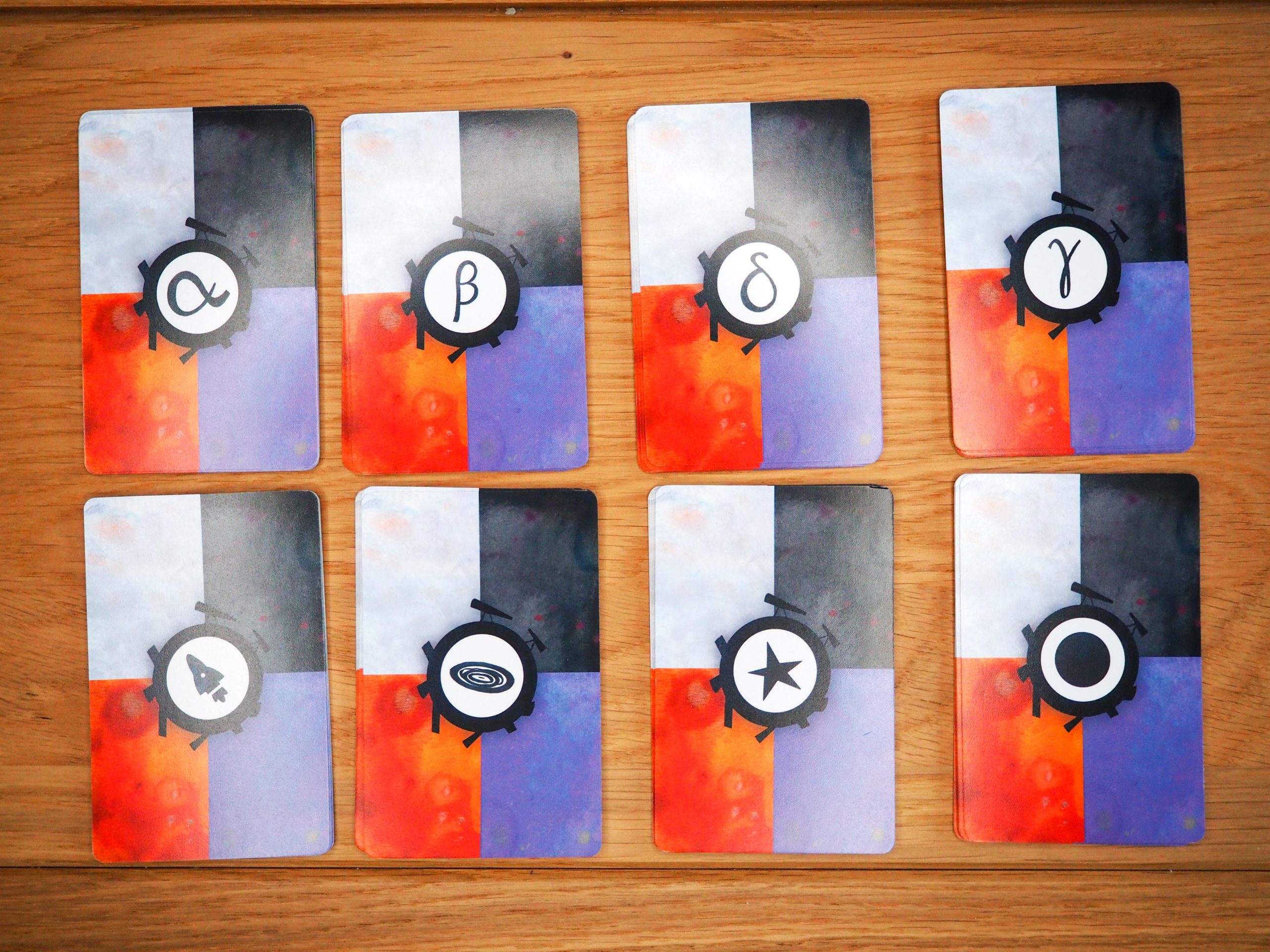 Auf dem Bild sind acht Kartenstapeln in zwei Reihen zu je vier Stapeln zu sehen. Jede Karte hat vier rechteckige farbige Bereiche. Weiß, schwarz, orange und lila. Mittig auf der Karte sind in Kreisen die Zeichen der Karten zu erkennen. 