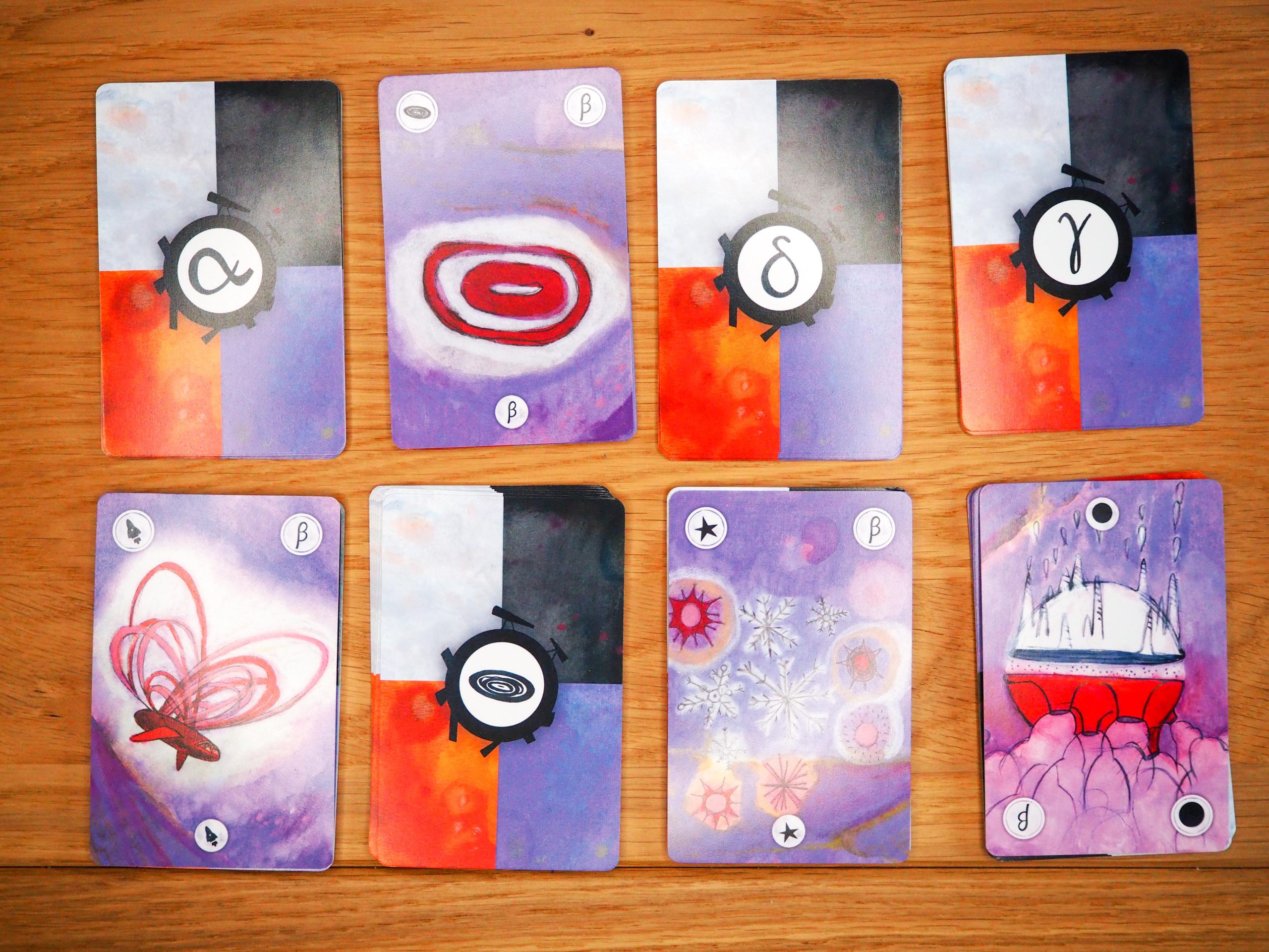 Auf dem Bild sind acht Kartenstapeln in zwei Reihen zu je vier Stapeln zu sehen. Jede Karte hat vier rechteckige farbige Bereiche. Weiß, schwarz, orange und lila. Mittig auf der Karte sind in Kreisen die Zeichen der Karten zu erkennen. Vier Karten sind liegen aufgedeckt auf dem Stapel und zeigen die gleiche Galaxie.