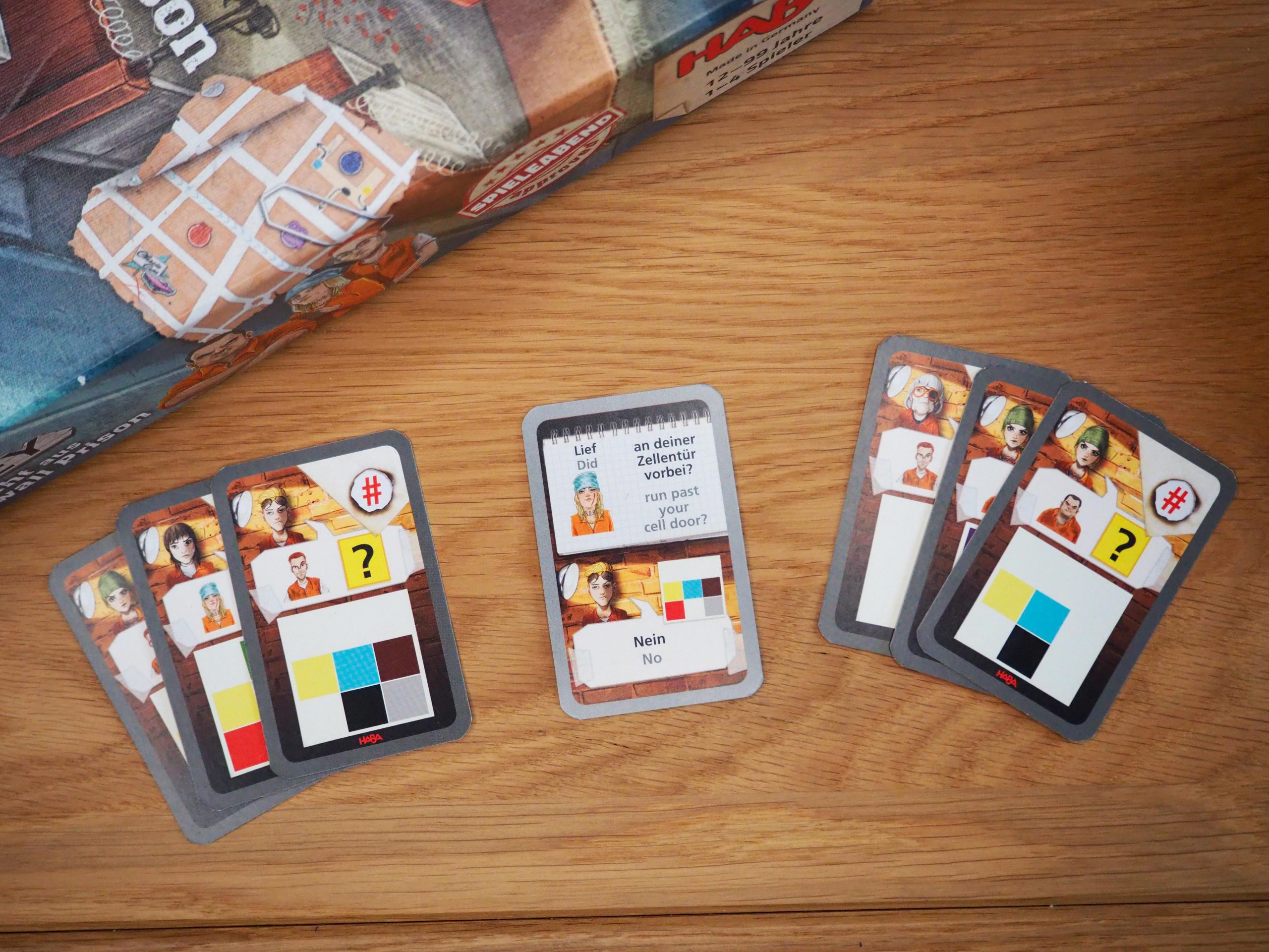 Links oben im Bild ist ein Teil der Spielschachtel zu sehen. Im Vordergrund liegt eine kleine Bandenkarte aufgedeckt in der Mitte und jeweils drei weitere aufeinander als Fächer links und rechts von der aufgedeckten Karte. 