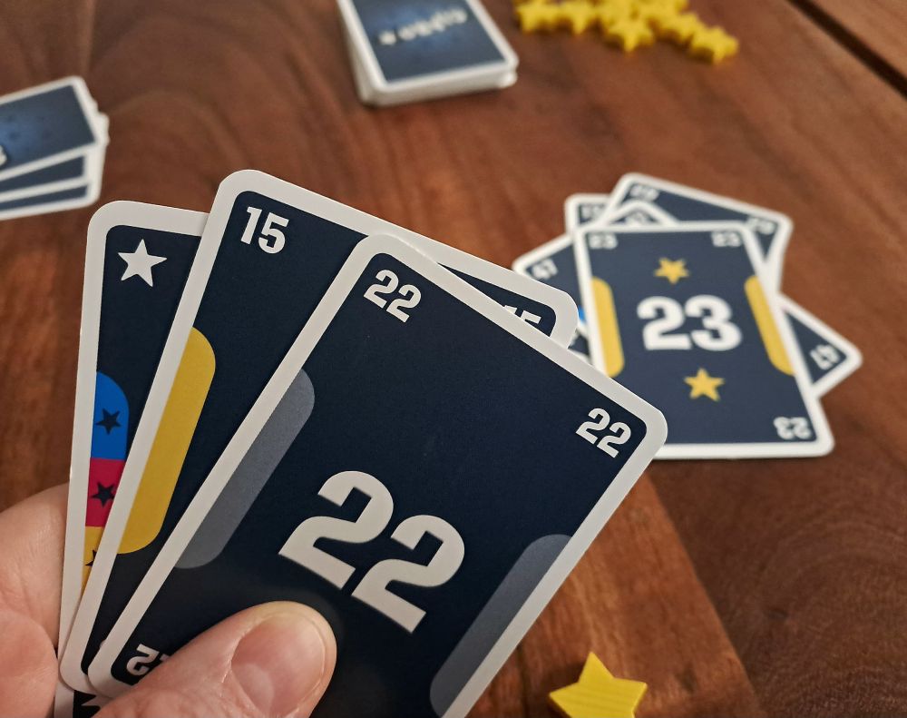 Eine Kartenhand mit einem Joker, einer 15 und einer 22. In der Tischmitte liegt eine 23