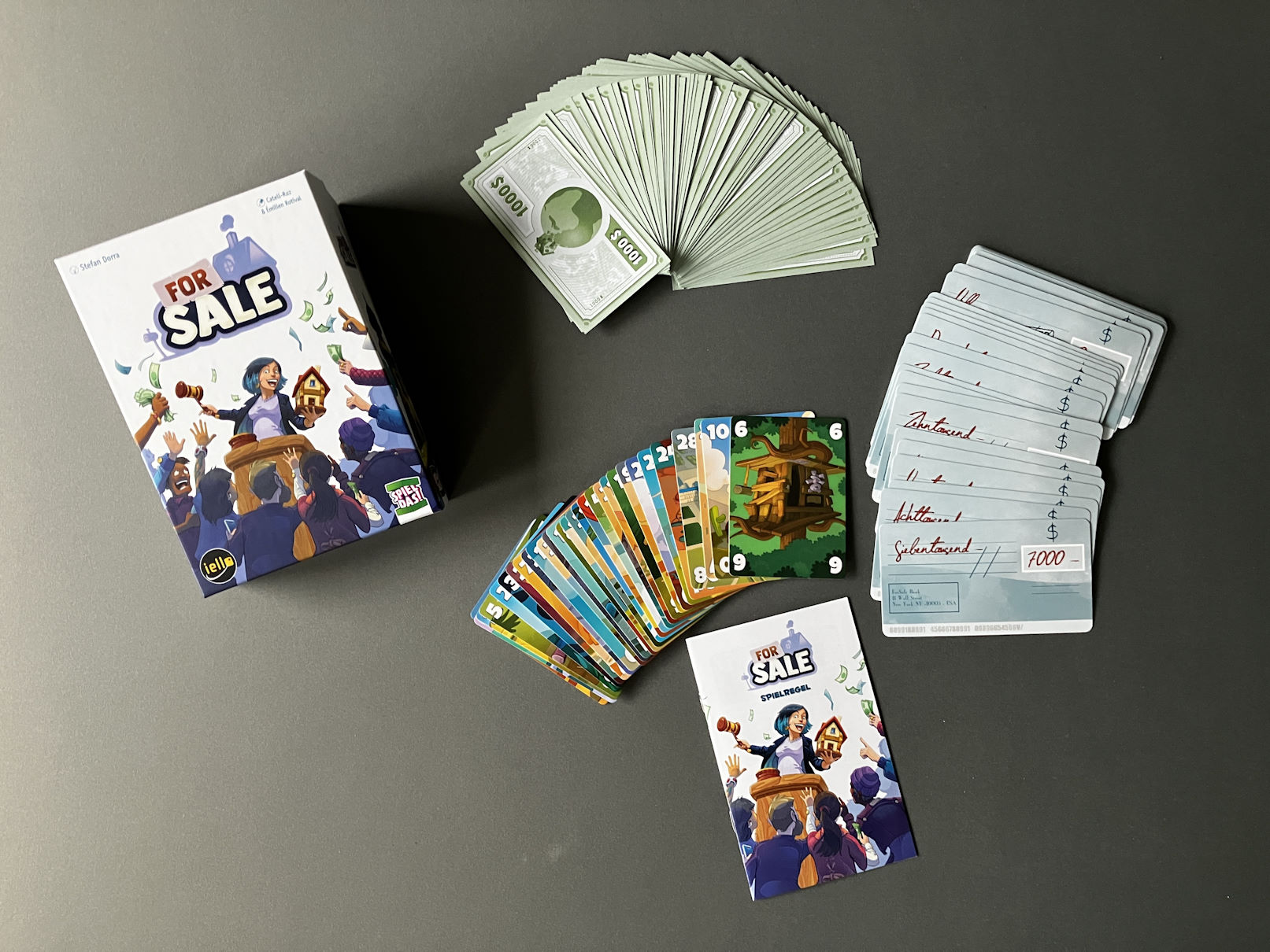 Die Inhalte des Spiels liegen auf dem Tisch aufgefächert: 84 Geldscheine, 30 Schecks und 30 Immobilienkarten.
