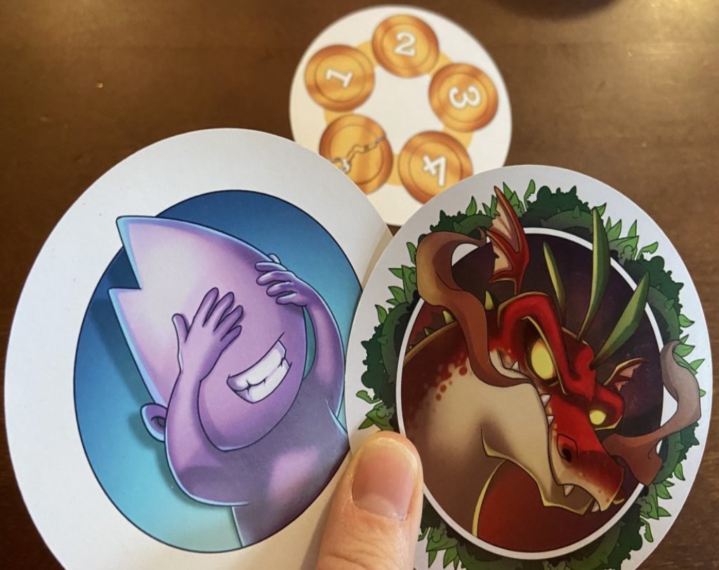 Hand mit einer Karten mit einem Drachen und einer Karte mit einem Männchen, dass sich die Augen zuhält. Im Hintergrund eine Karte mit Münzen.