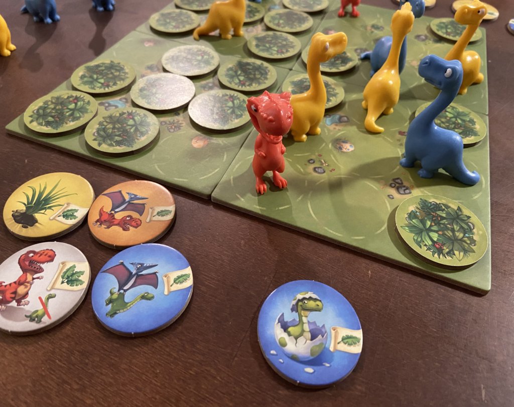 Spielplan mit Dinos und Plättchen, daneben liegen verschiedene Plättchen offen, auf dem vorderen ist ein Dino der aus einem Ei schlüpft zu sehen.