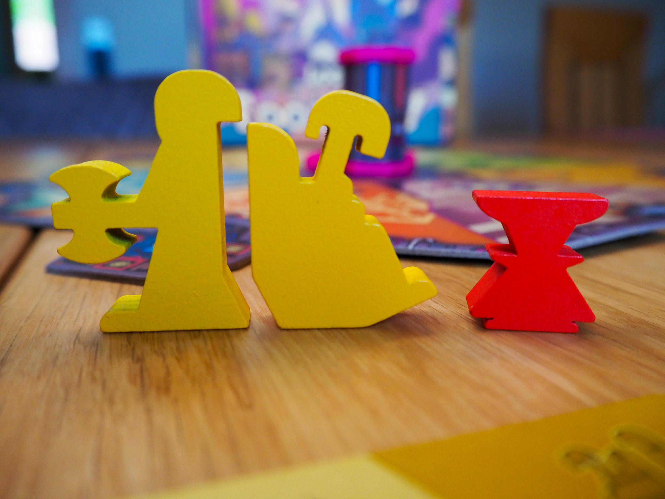 Im Hintergrund erscheint unscharf das aufgebaute Spielbrett inklusive der Spielschachtel der Erweiterung. Im Vordergrund stehen die Spielfiguren der neuen Charaktere. Die Zwillingen sind links angeordnet und erscheinen in knalligem gelb wie ihre Charakterkarte. Die halb so große Spielfigur des Foo Klein Schlags hingegen ist knallig rot.