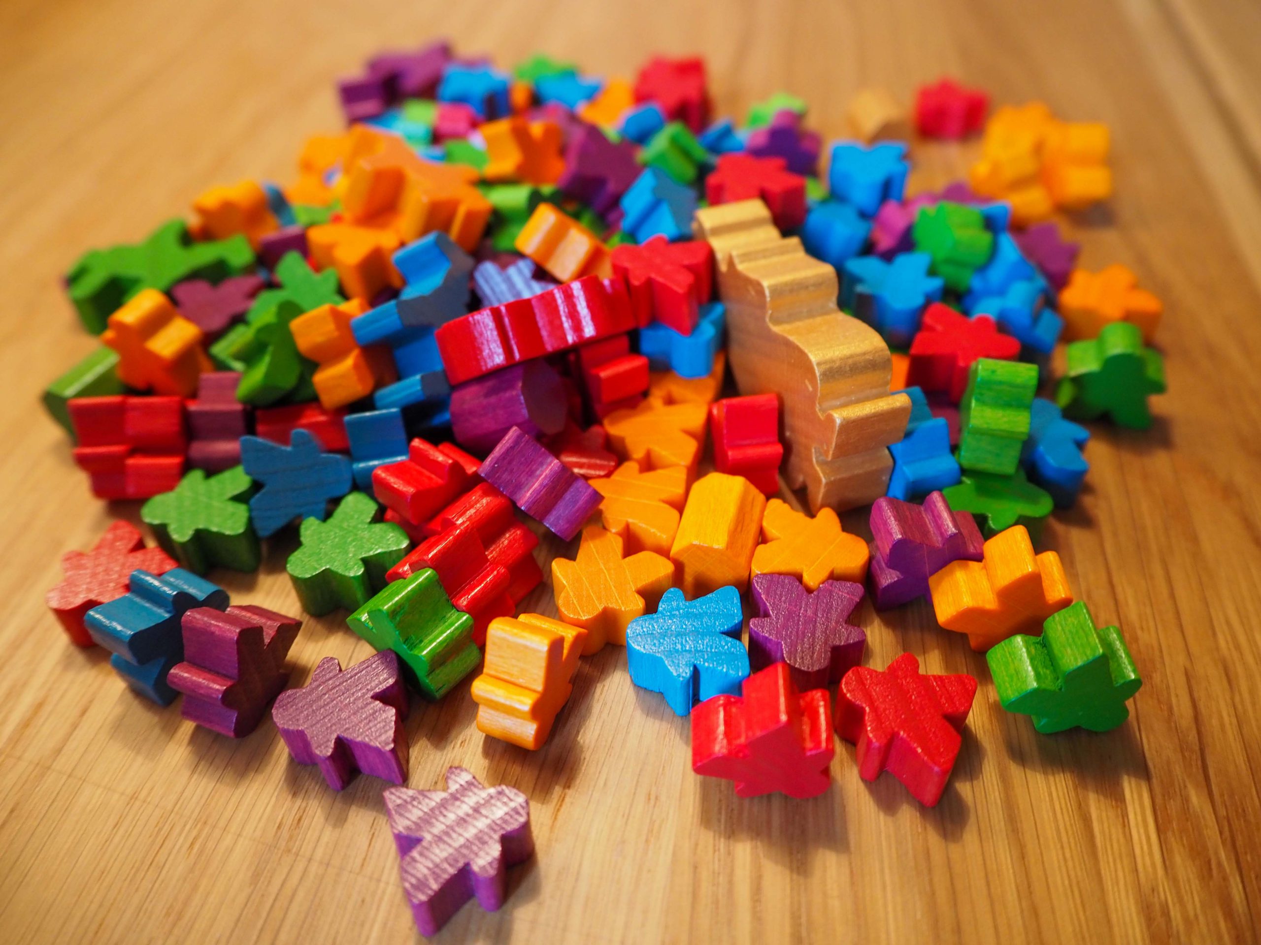 Auf einem mittelbraunen Holztisch ist ein Haufen aus Spielfiguren zu sehen. Die Spielfiguren haben die Farben lila, grün, gelb, blau und rot. Sie Farben sind kräftig und glänzend. Etwas rechts der Mitte liegt eine größere, goldene Spielfigur auf der Seite. Diese stellt den König dar.