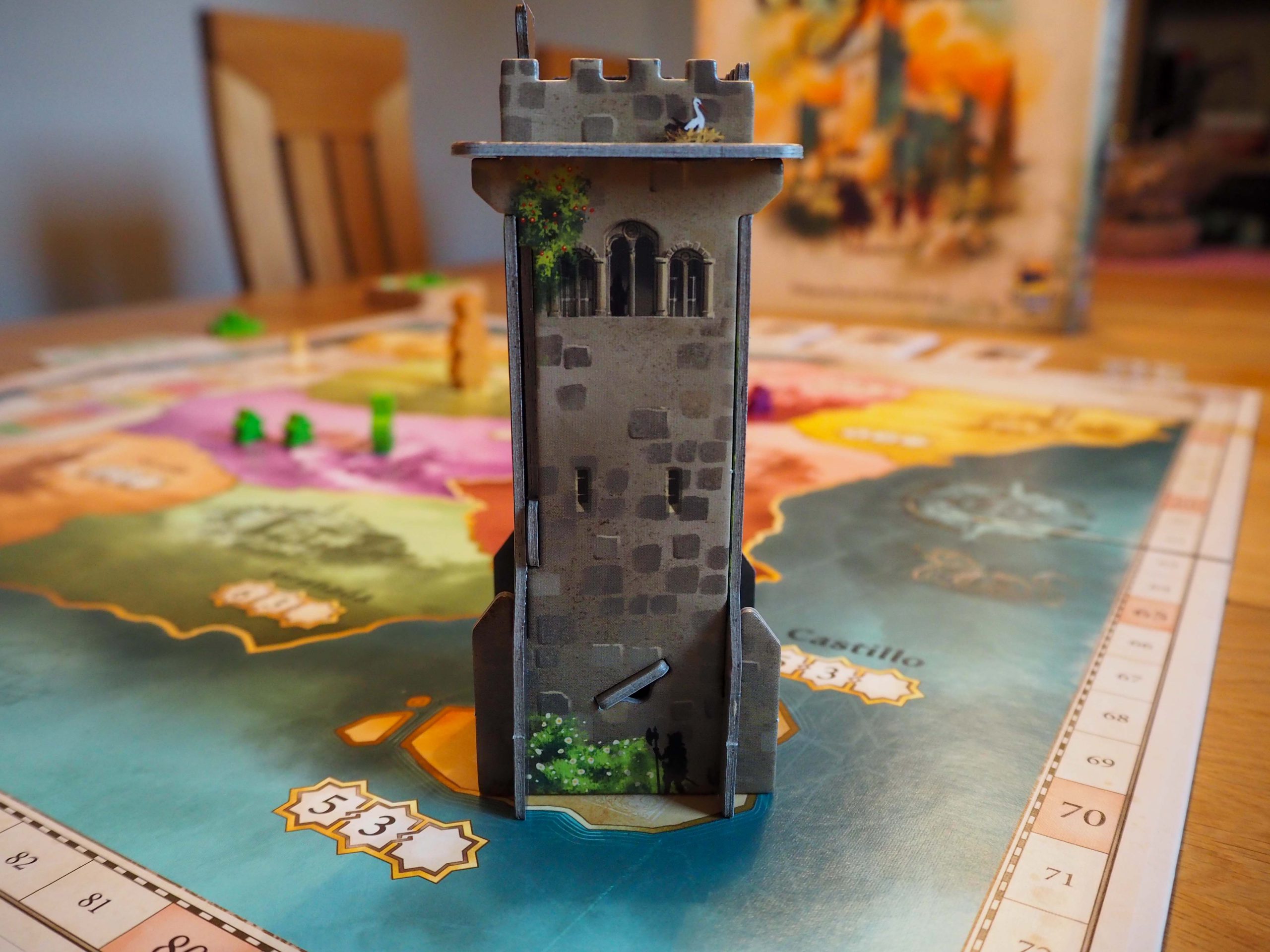 Mittig im Bild auf dem Spielplan steht der Castillo-Turm. Er hat Mauern in grau, braun und im oberen Drittel Turmzacken und drei angedeutete Fenster. Im Hintergrund ist unscharf die Spielepackung zu sehen.