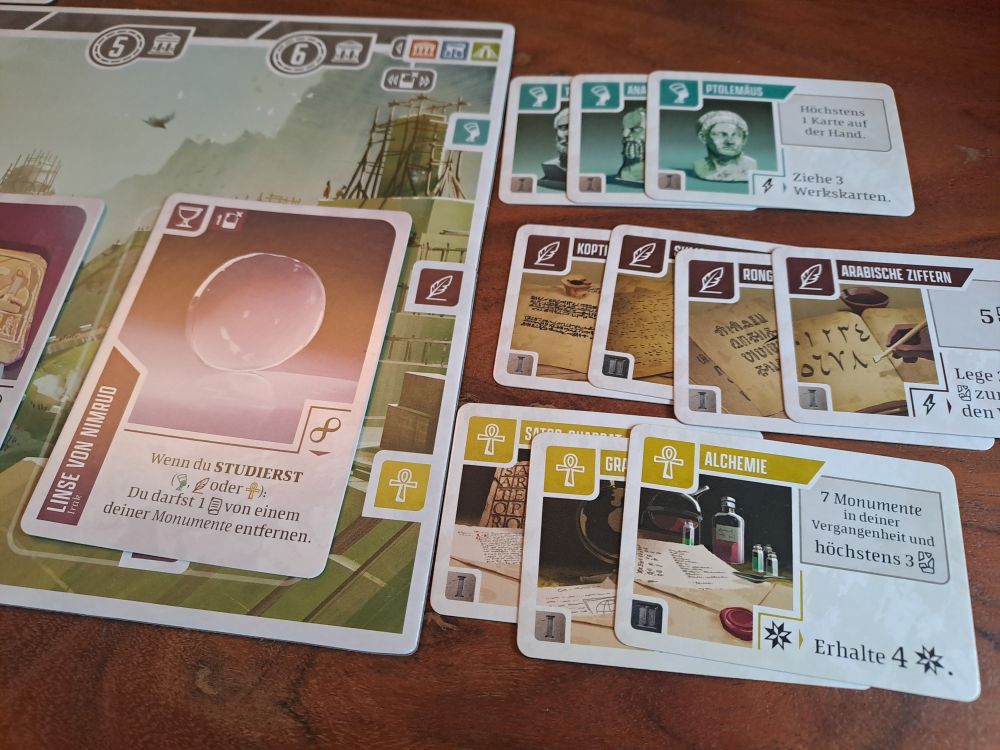 Zu sehen ist ein Spieltableau an das bereits einige Erkenntniskarten angelegt wurden. 