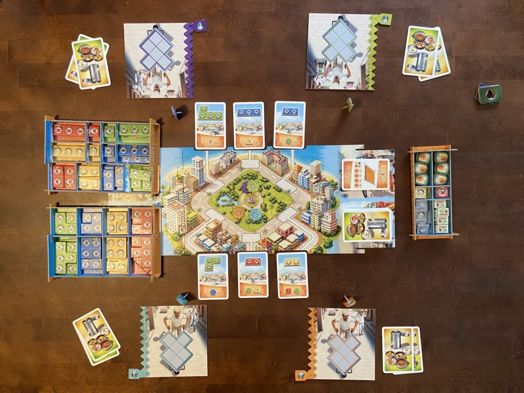 Zu sehen sind das Spielbrett mit Karten drumherum, sowie 4 Spieltableaus mit je zwei Karten