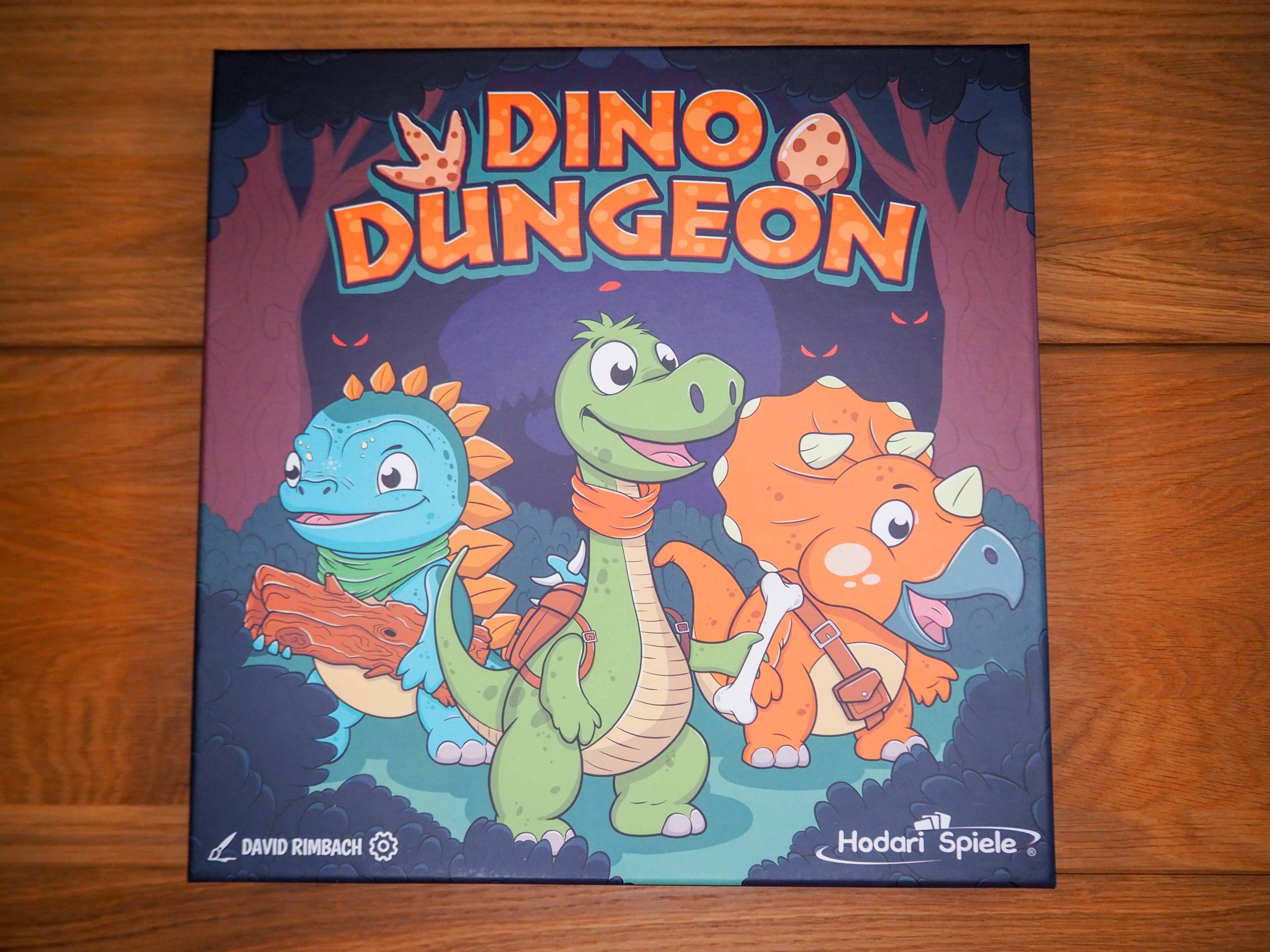 Schachtel auf Holztisch mit prominentem Titel in Farbe Orange. Darunter drei Dinos.