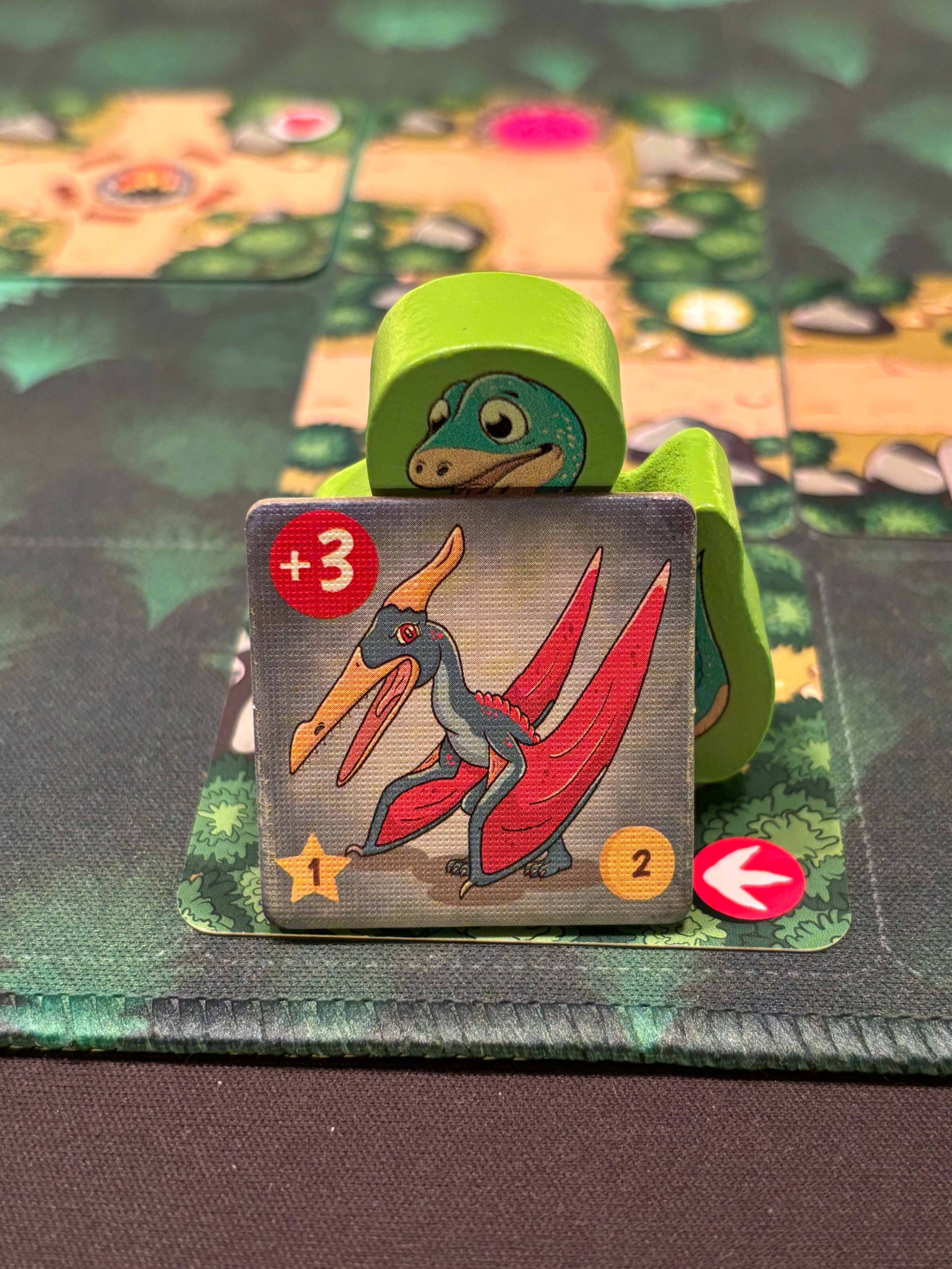 Grüne Dino Figur aufrecht stehend und eine rechteckige Kachel mit Bösling davor.