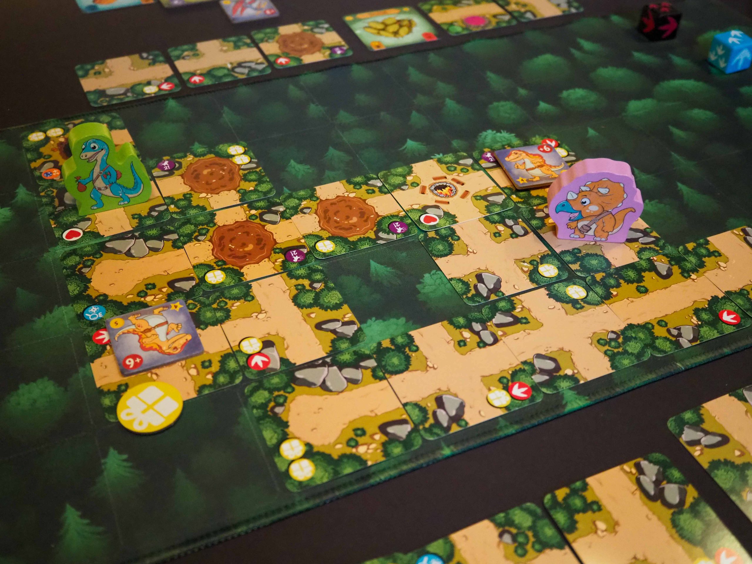 Auf grünem Untergrund viele ausgelegte quadratische Entdeckungskarten mit verschachtelten Wegen. Darauf an verschiedenen stellen die Dino Holzfiguren.