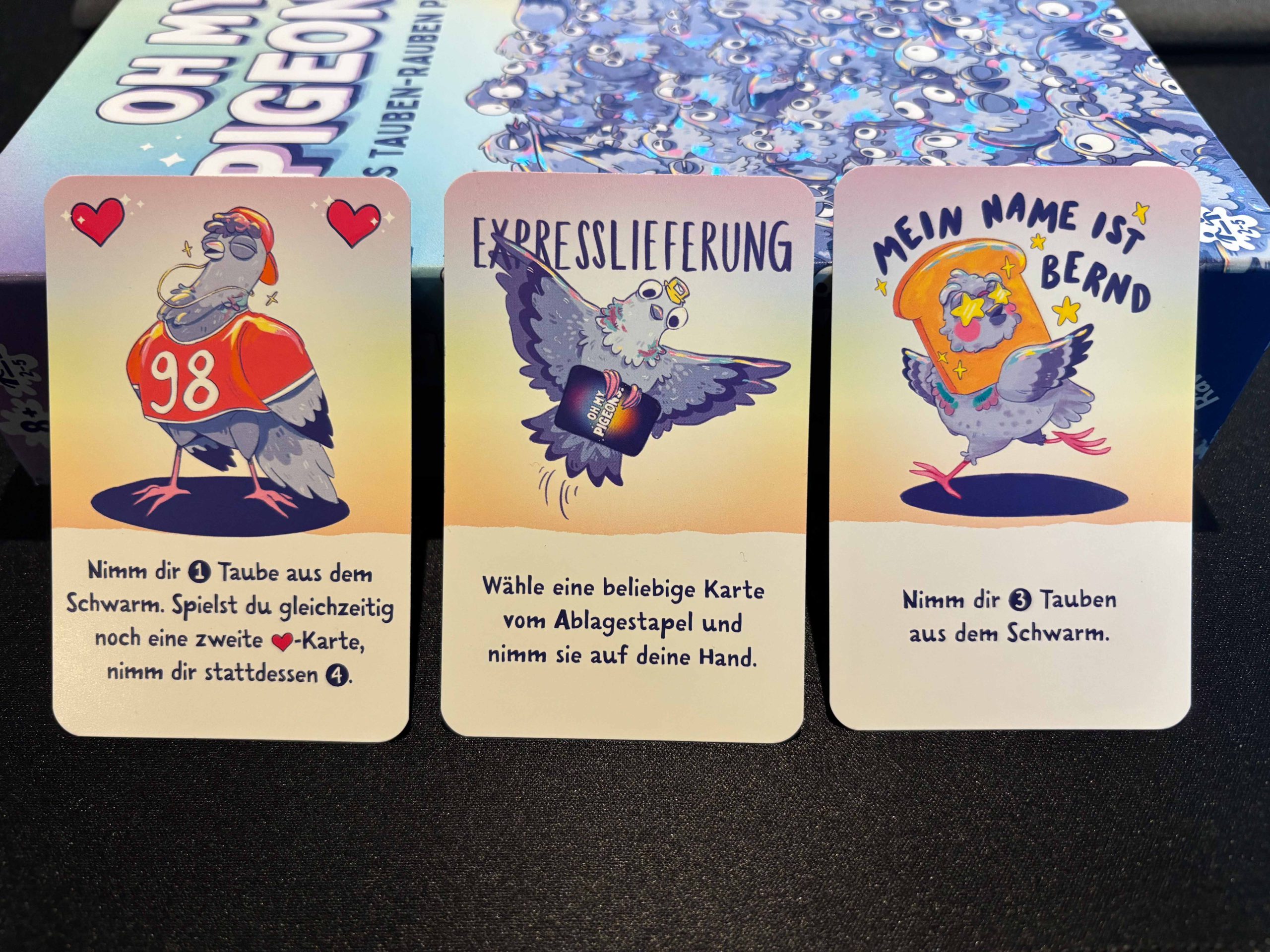 Drei aufrecht stehende Spielkarten angelehnt am liegenden Spielkarton.