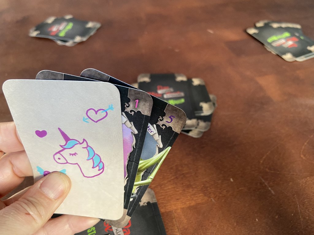 Zu sehen sind 3 Handkarten, die mit einer Lüge abgelegt werden (möchten). Im Hintergrund ist der verdeckte Ablagestapel zu sehen.