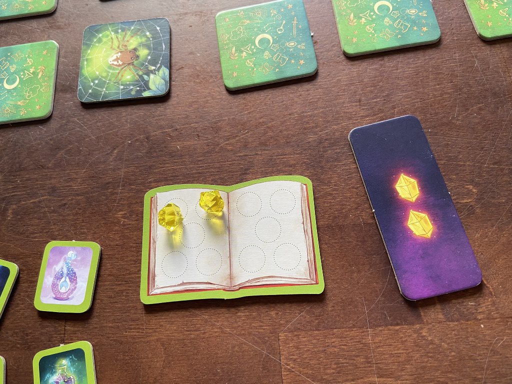 Ein erfülltes Aufgabenplättchen zeigt zwei Kristalle, die auf das eigene Buch gelegt werden.