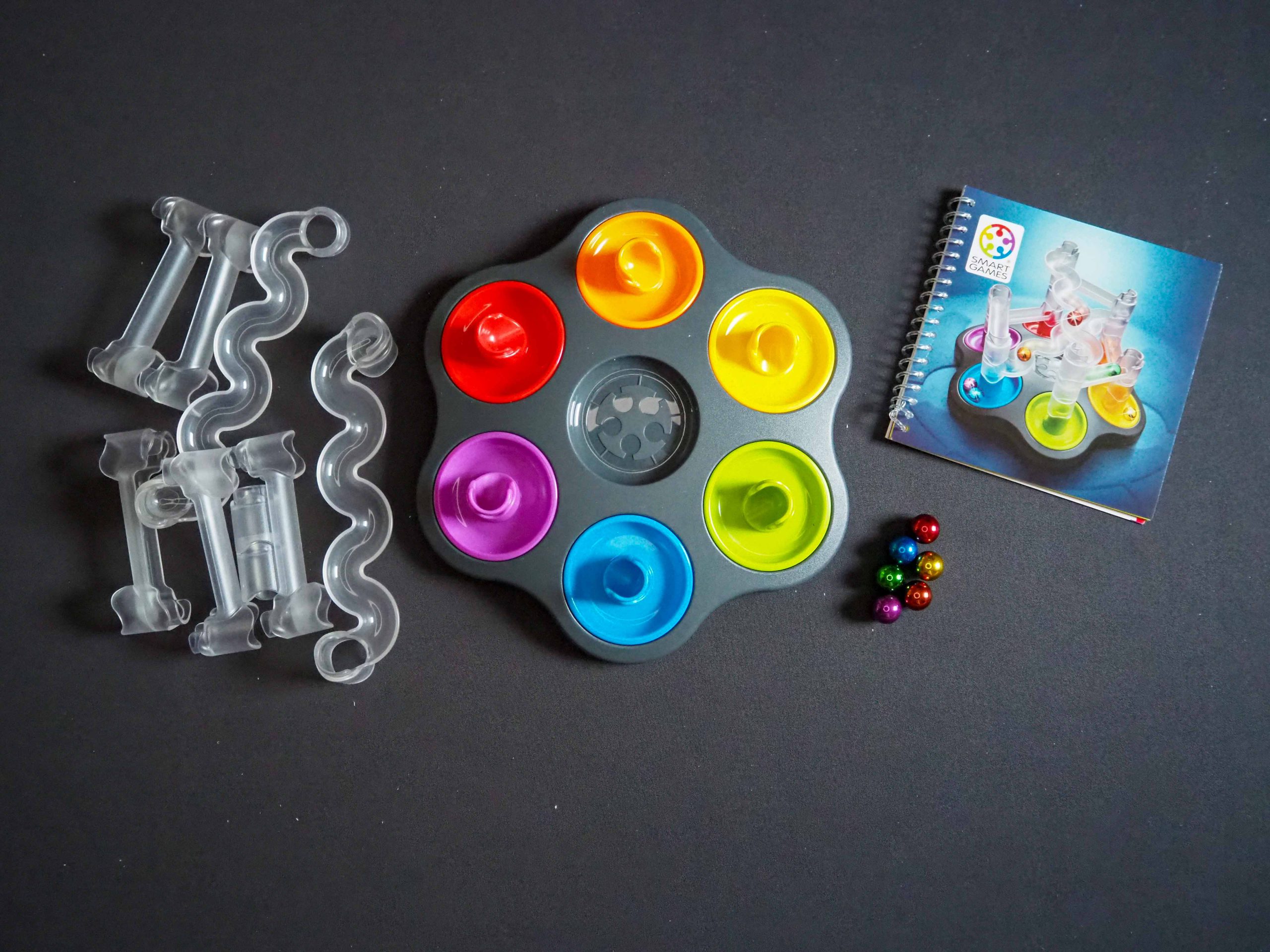 Spielmaterial liegt verteilt. Links durchsichtige Streckenabschnitte, mittig das Spielfeld rund mit verschiedenfarbigen Zonen und rechts sechs Kugeln verschiedener Farben und ein Ringbuch mit den Aufgaben.
