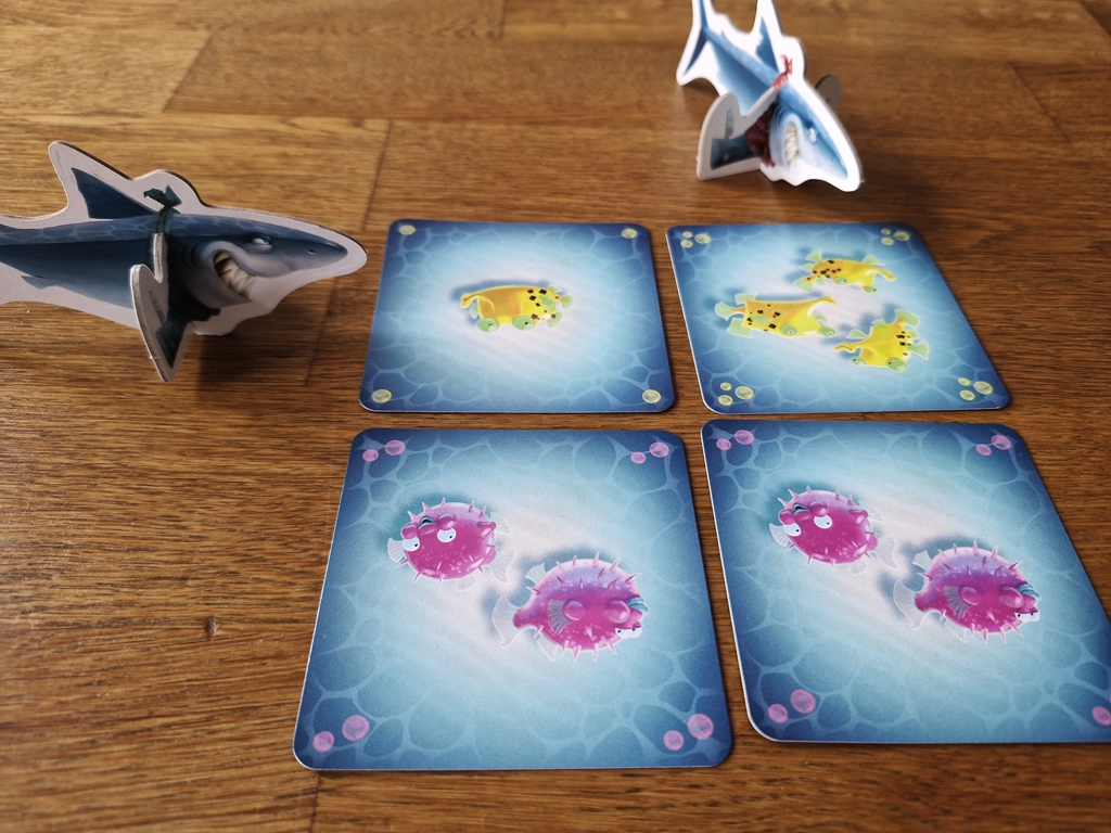 Vier ausliegende Karten, zwei Haifiguren markieren Reihe und Spalte mit Mehrheit an Fischen