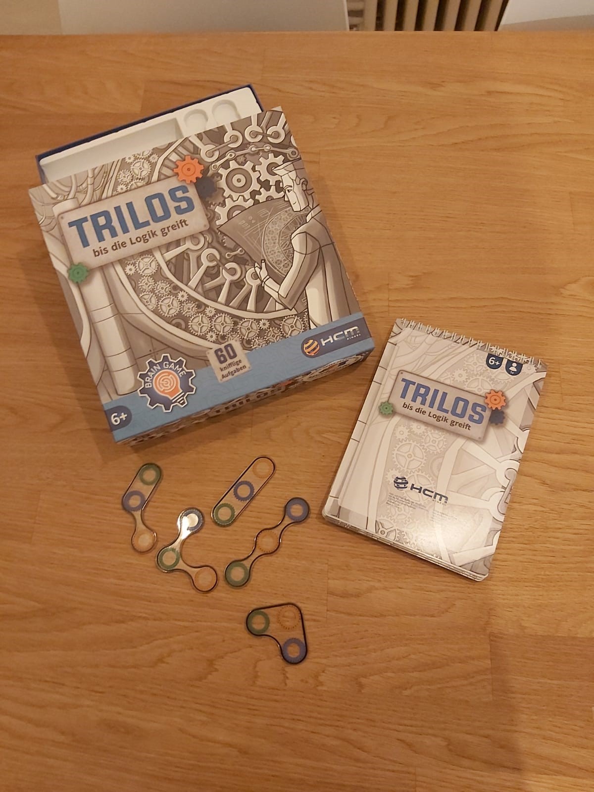Zu sehen sind die Verpackung des Spiels "Trilos" sowie der Aufgabenblock und die 5 Zahnrad-Aufleger unterschiedlicher Formen.
