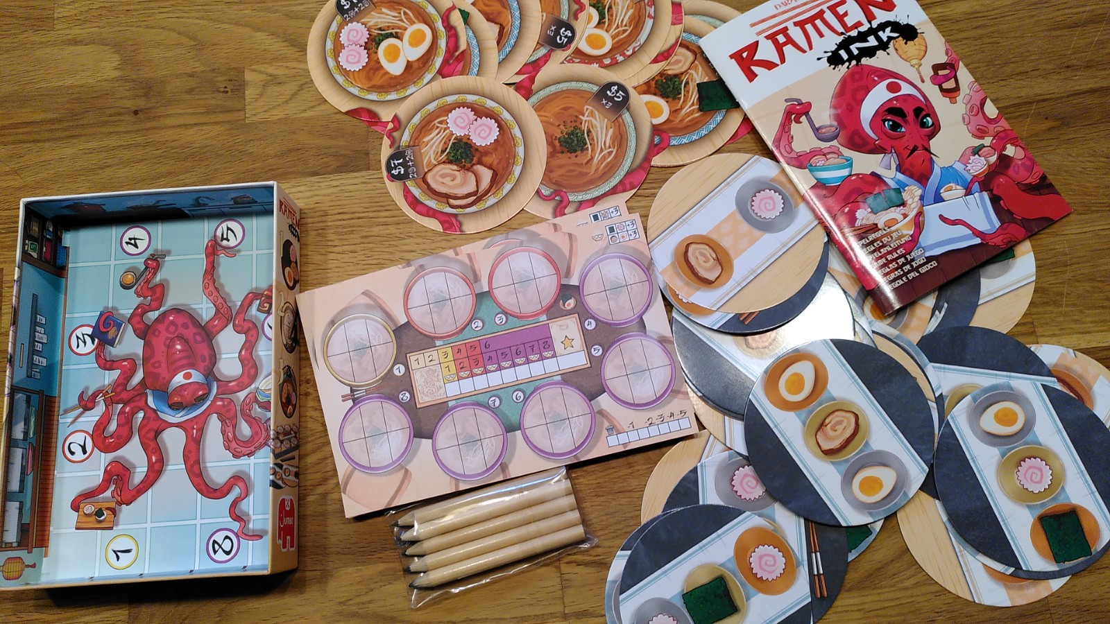Spielmaterial von Ramen Ink, runde Karten mit Zutaten und Suppenportionen, Spielblock und der Oktopus-Koch auf dem Schachtelboden