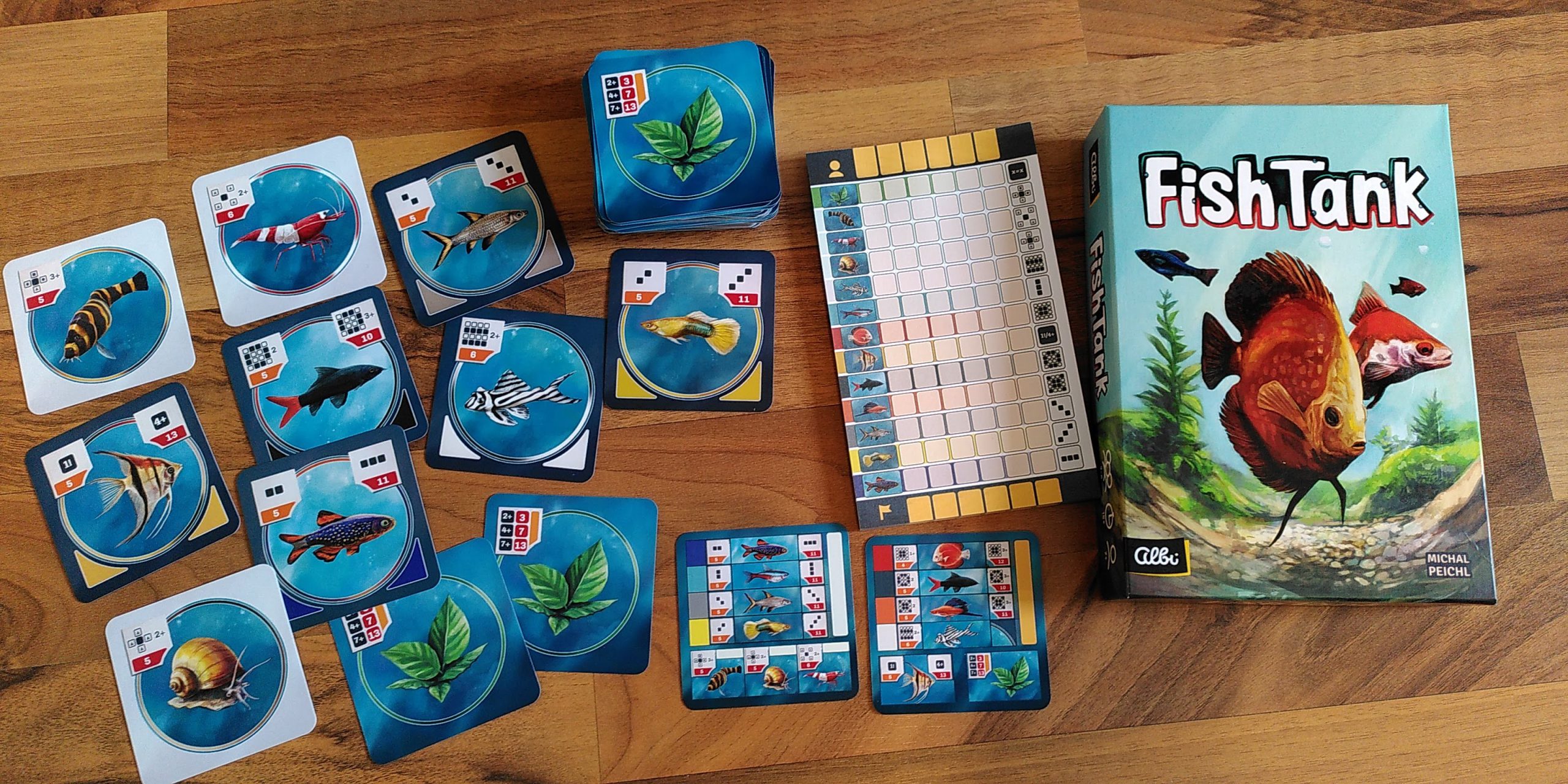 Übersicht über das Spielmaterial: quadratische Fisch- und andere Tierkarten, auf der Rückseite Wasserpflanzen, Übersichtskarten und Punkteblock