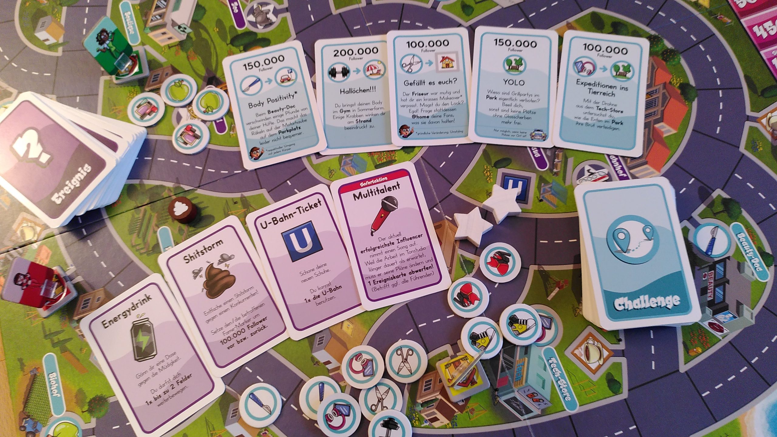 Überblick über das bunte lustige Spielmaterial, Ereigniskarten, Challenge-Karten, bunte Ortstoken, der Stadtplan als Spielplan und unsere Influencer-Spielfiguren