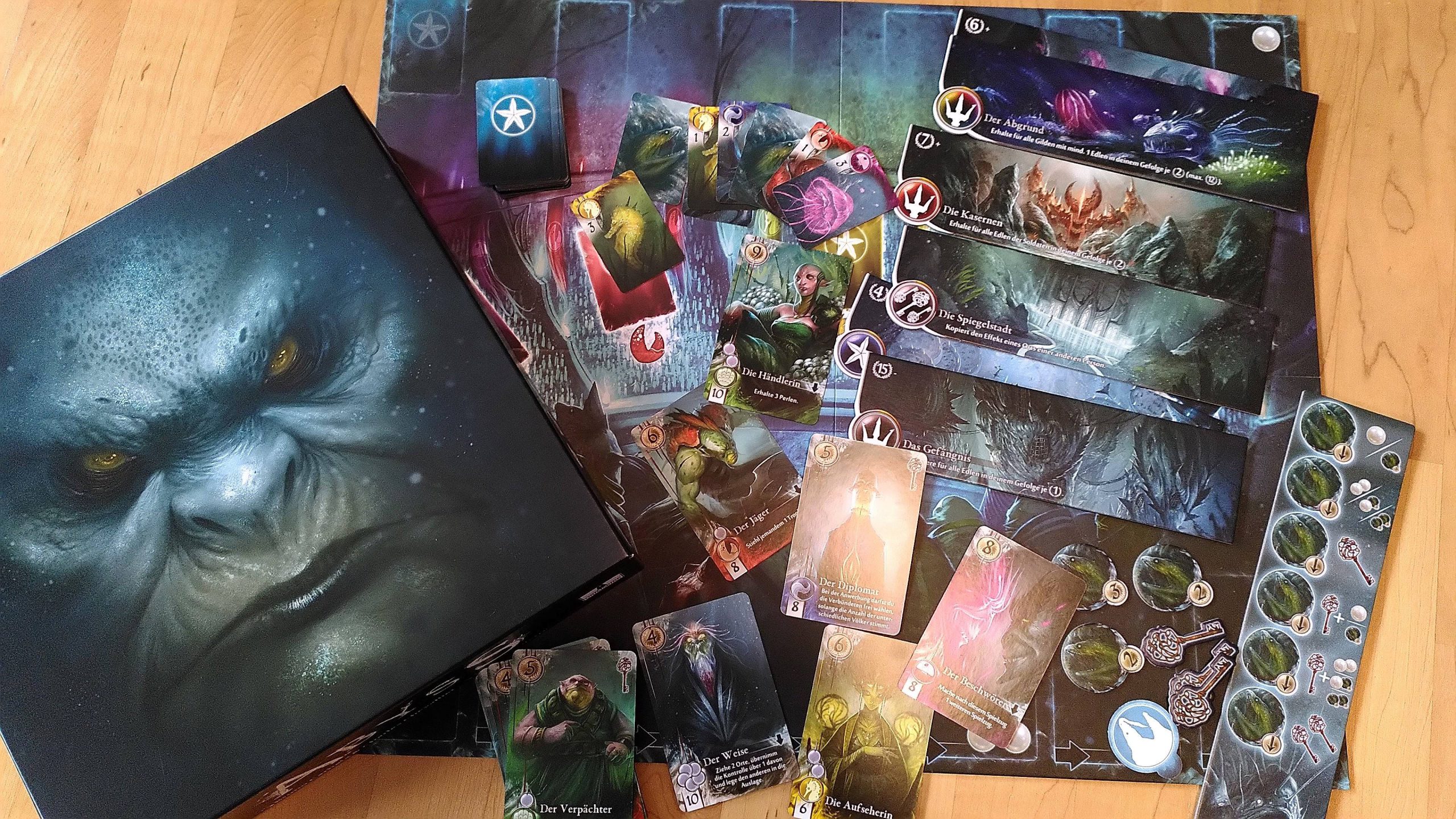 Überblick über das Spielmaterial und das Design von Abyss. Die imposante Schachtel, viele Karten mit Unterwasserwesen, Papp-Schlüssel und -Abzeichen