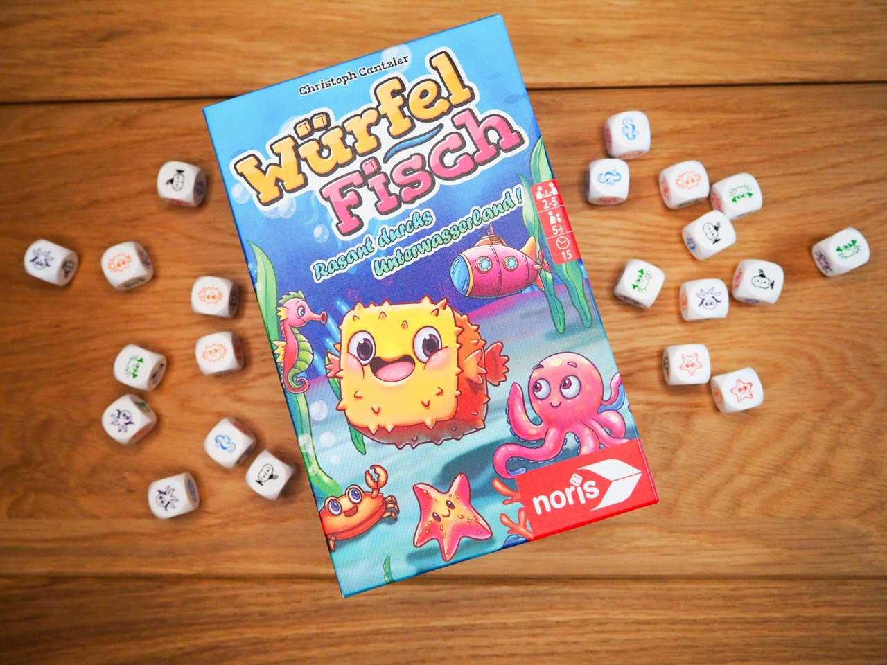 Fröhlich schaut uns ein gelber Kugelfisch von der Spielebox aus an. Die Spielebox liegt auf einem Holztisch schräg mittig. Links und rechts neben der Spielebox liegen verstreut ein paar Würfel mit unterschiedlichen Tierikonografien.