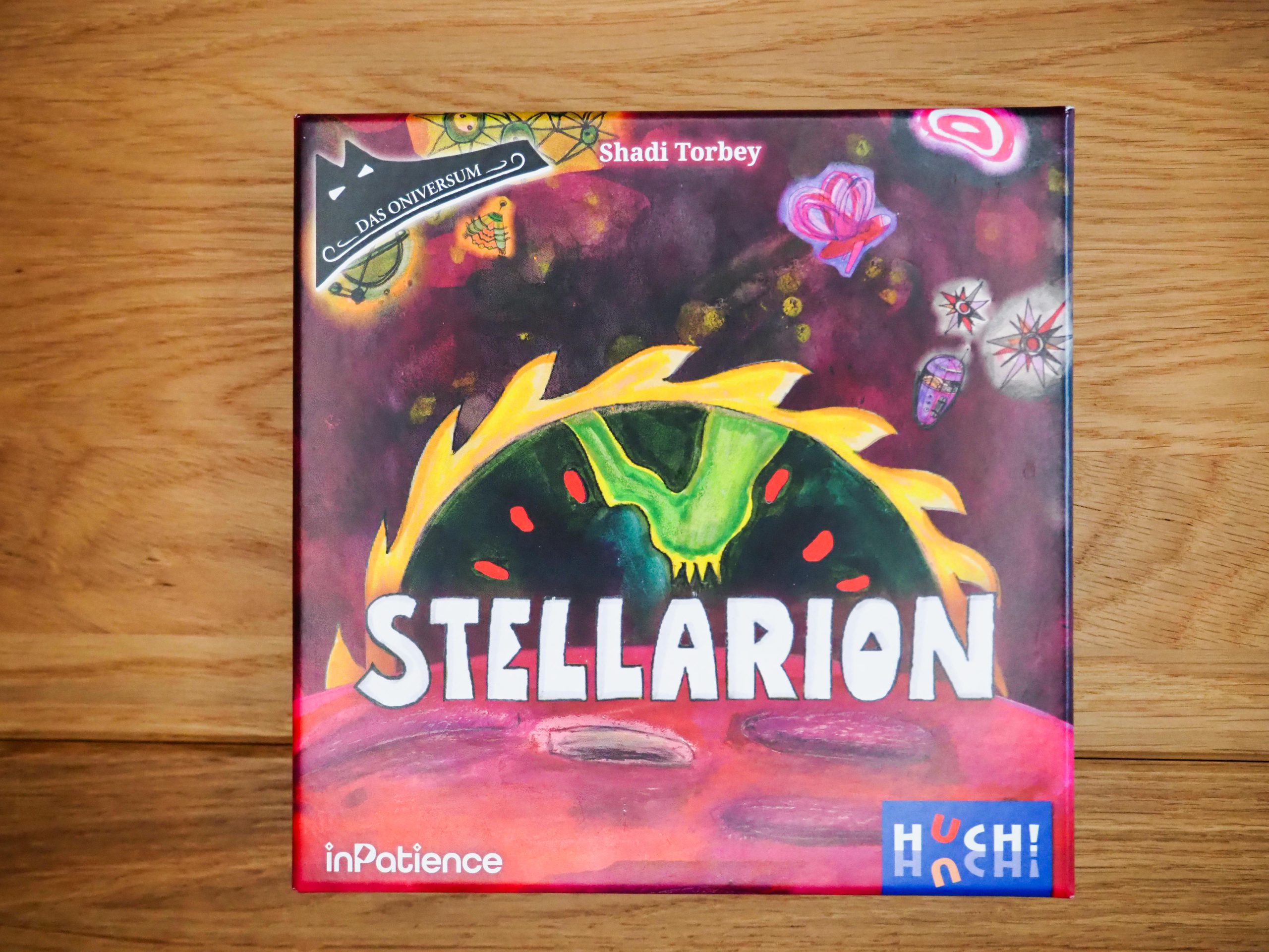 Auf einem Eicheholztisch liegt die Spielebox mittig im Bild. Die Schachtel ist in rosa/pink gehalten. Mit weißer, dick gedruckter Schrift erscheint der Titel in Großbuchstaben im unten Drittel der Schachtel. Eingerahmt wird die Überschrift von einem gelben Halbkreis mit Zacken.